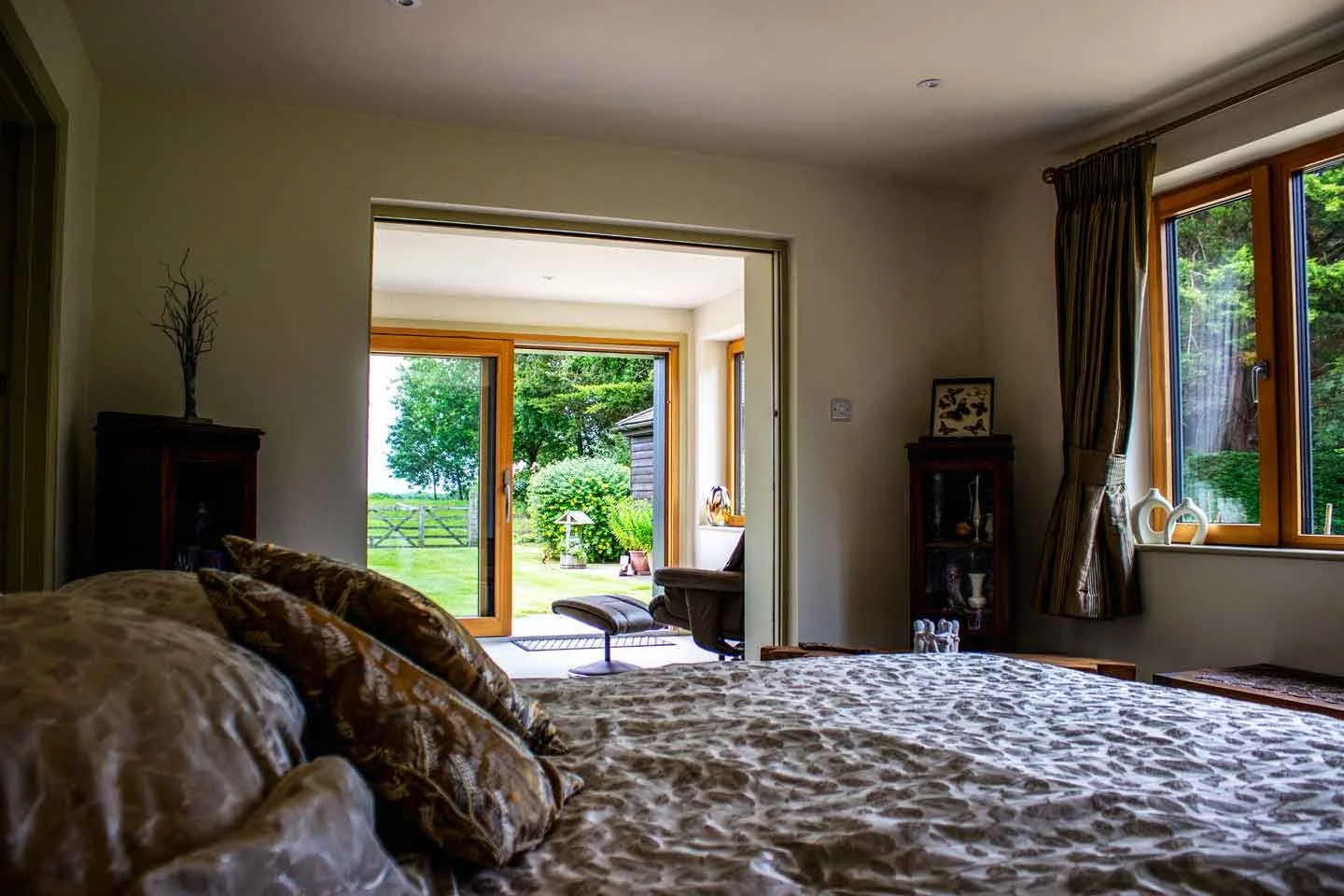 View from a bedroom with a bed in the foreground, overlooking a sitting area outside with green grass, trees, and a wooden fence.