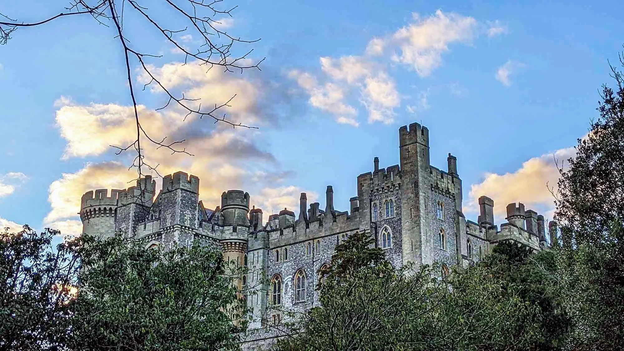 A historic castle with stone walls and battlements, surrounded by trees and under a partly cloudy sky during sunset.