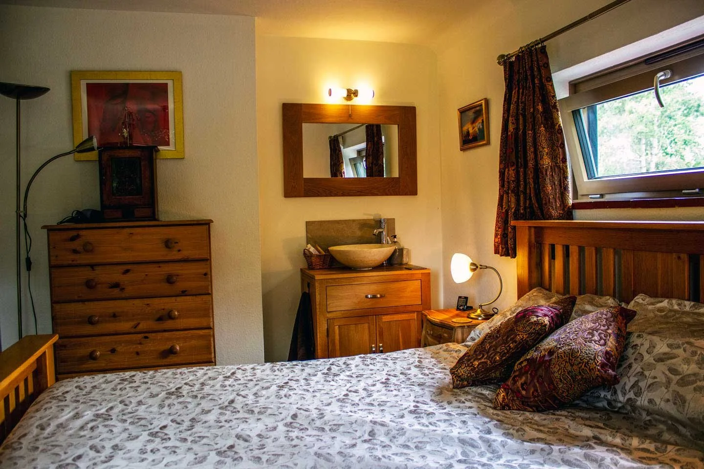 A cozy bedroom with a wooden bed frame, decorative pillows, a small bedside lamp, a dresser, and a sink with a mirror, framed pictures on the wall, and a window with a curtain.