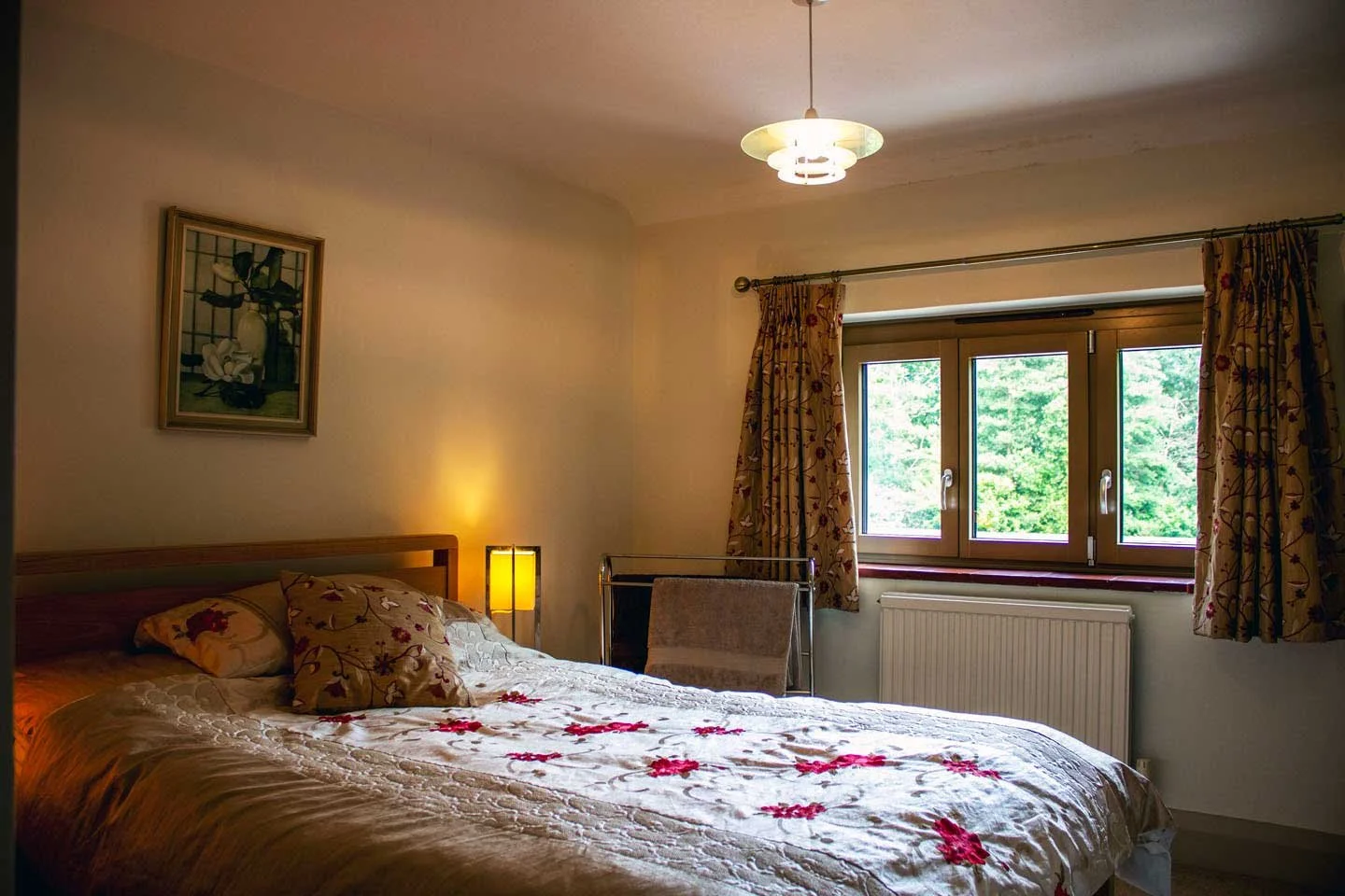 A bedroom with a bed, floral bedding, a bedside lamp, a window with floral curtains, and a painting on the wall.