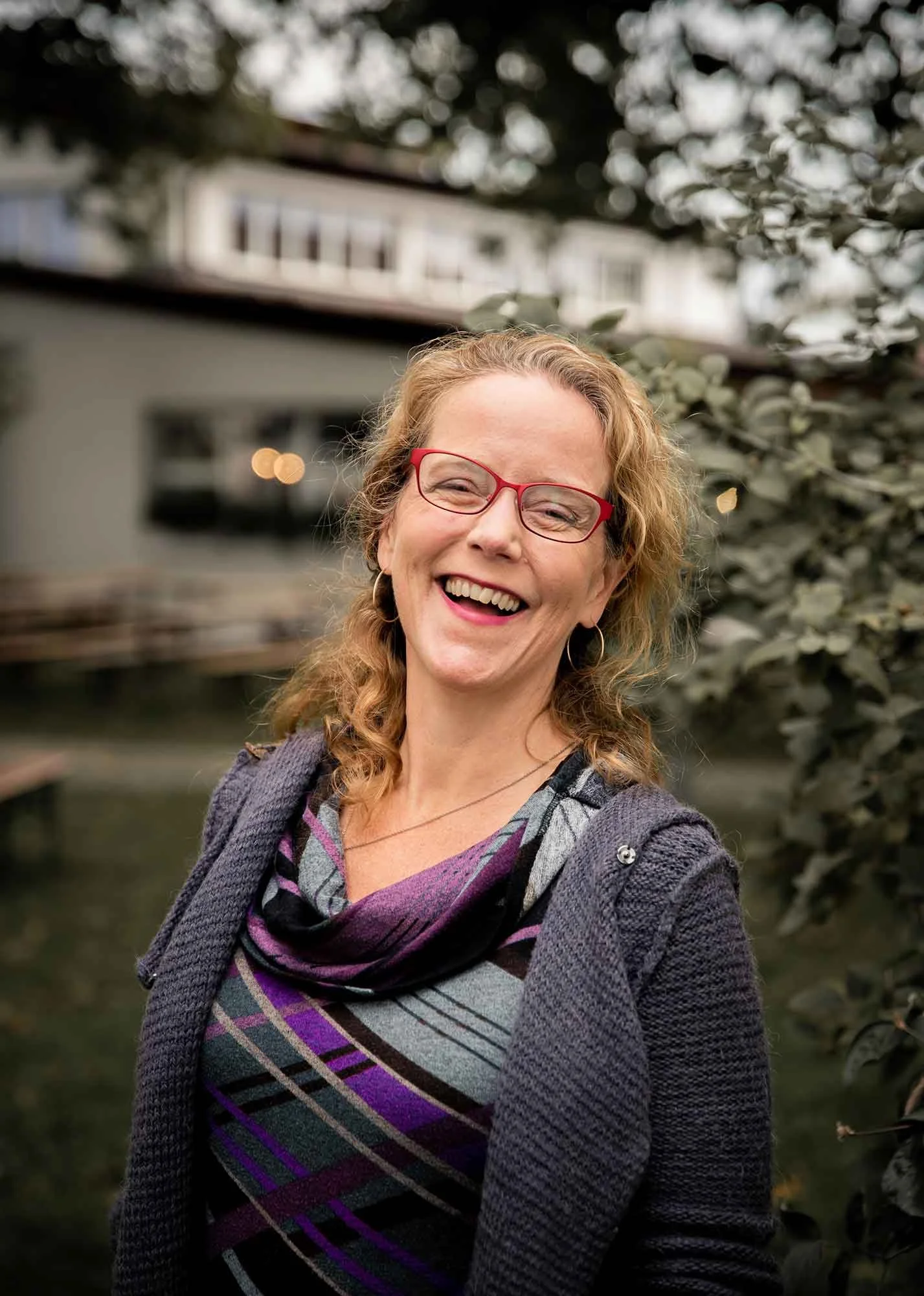 A smiling woman with curly red hair, red glasses, a colorful scarf, and a gray knitted cardigan standing outdoors with green foliage and a blurred building in the background.