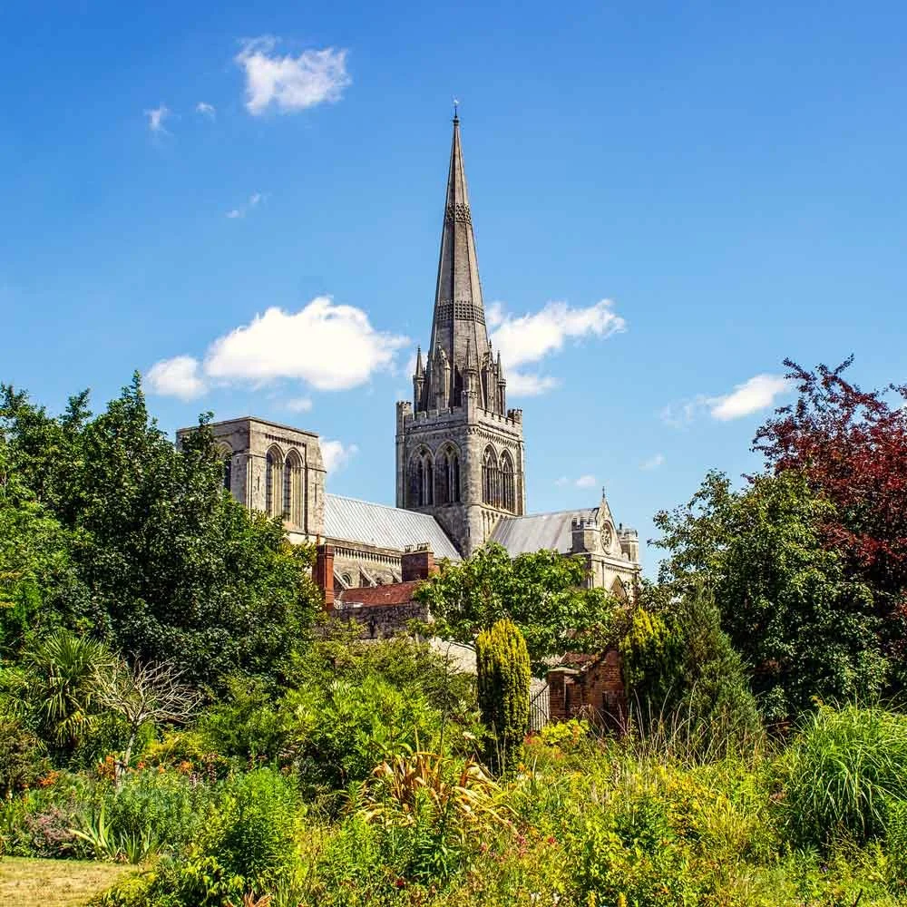 A historic stone church with a tall steeple, surrounded by green trees and a garden with colorful blooming plants, under a clear blue sky.