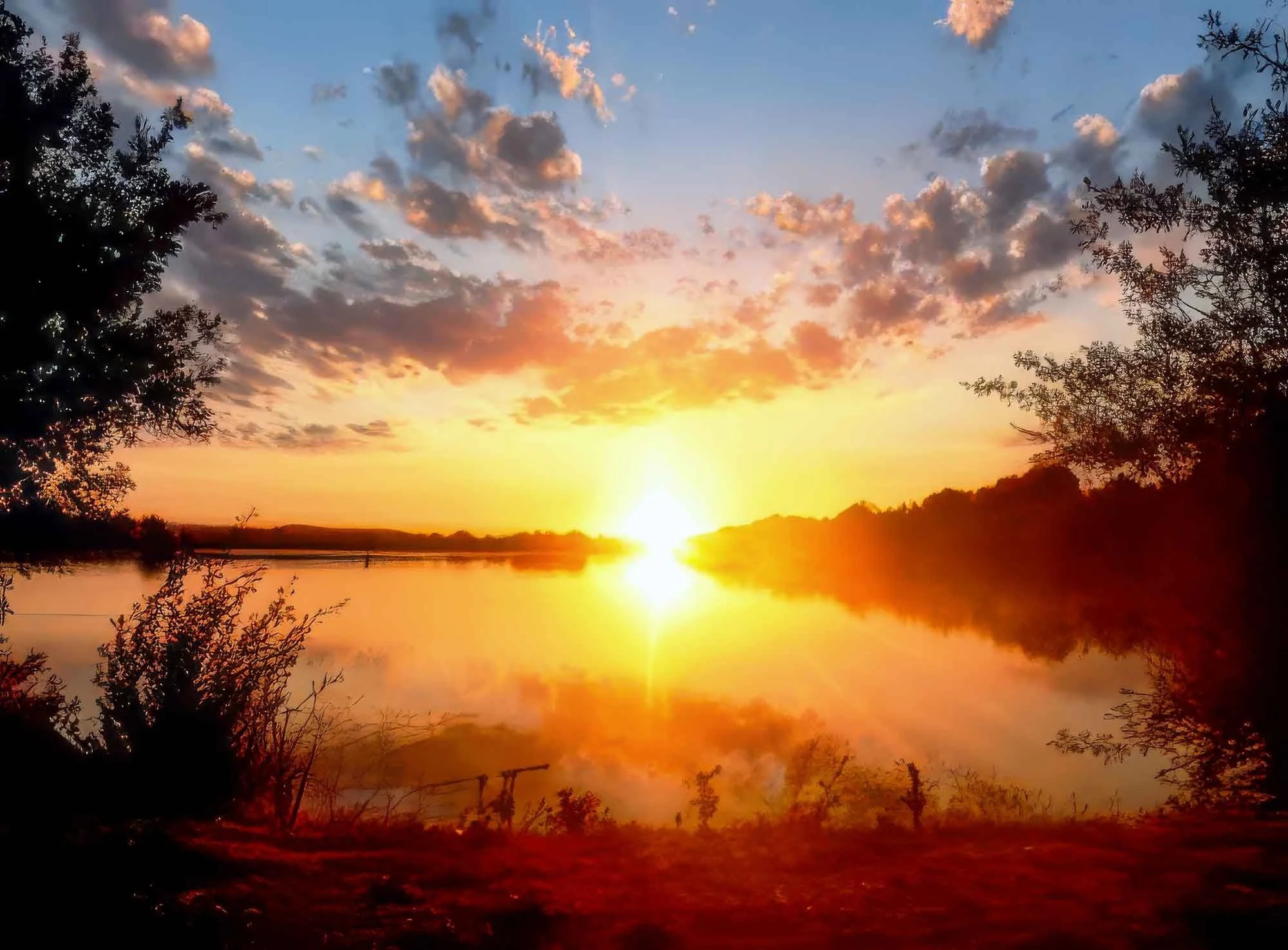 A sunset over a lake, with the sky filled with clouds illuminated in shades of pink, orange, and yellow. The sun is near the horizon, reflecting on the water, and silhouetted trees frame the scene.