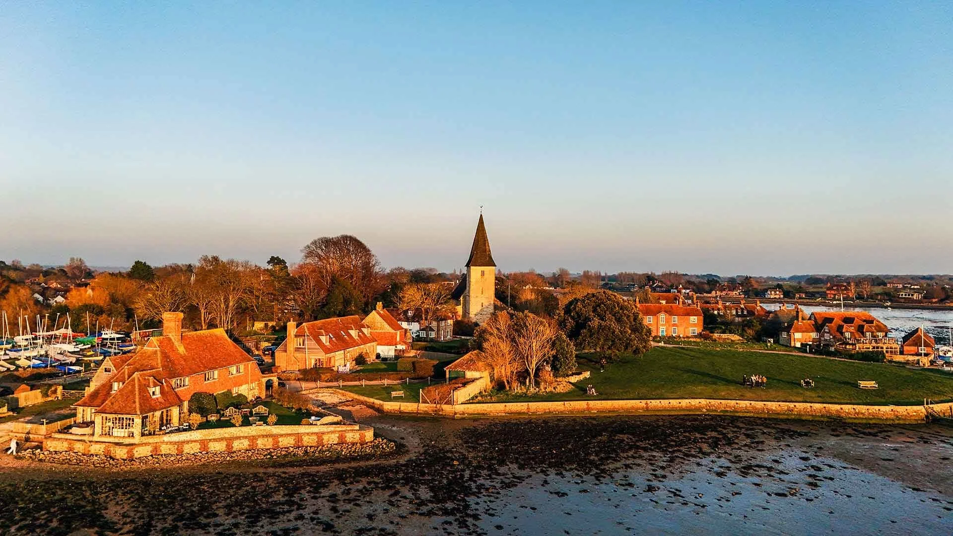 A coastal village during sunset with a church steeple, small houses, boats docked in a marina, and people and benches near the waterfront.