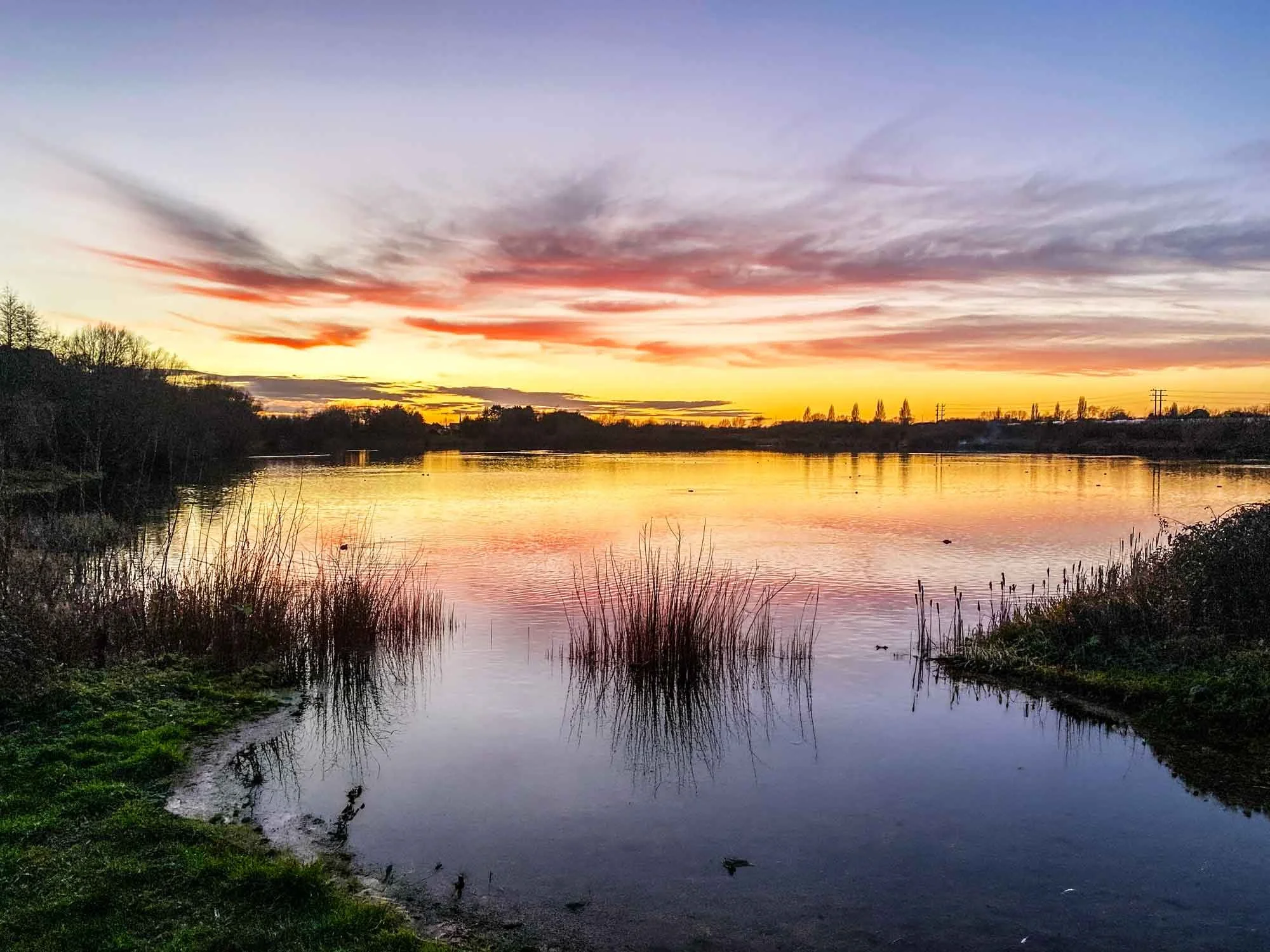 Sunset over a calm river with trees and bushes along the riverbank, reflecting the vibrant colors of the sky.