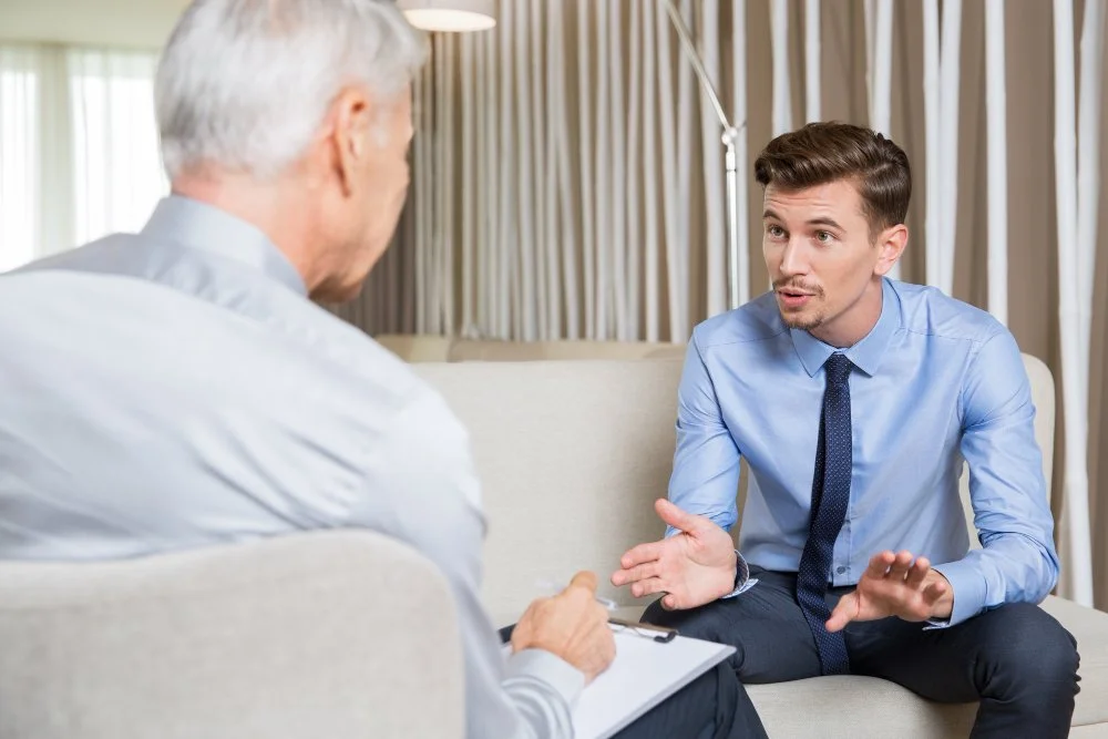 Young man talking to an older man in a professional setting, sitting on a sofa, with curtains in the background.