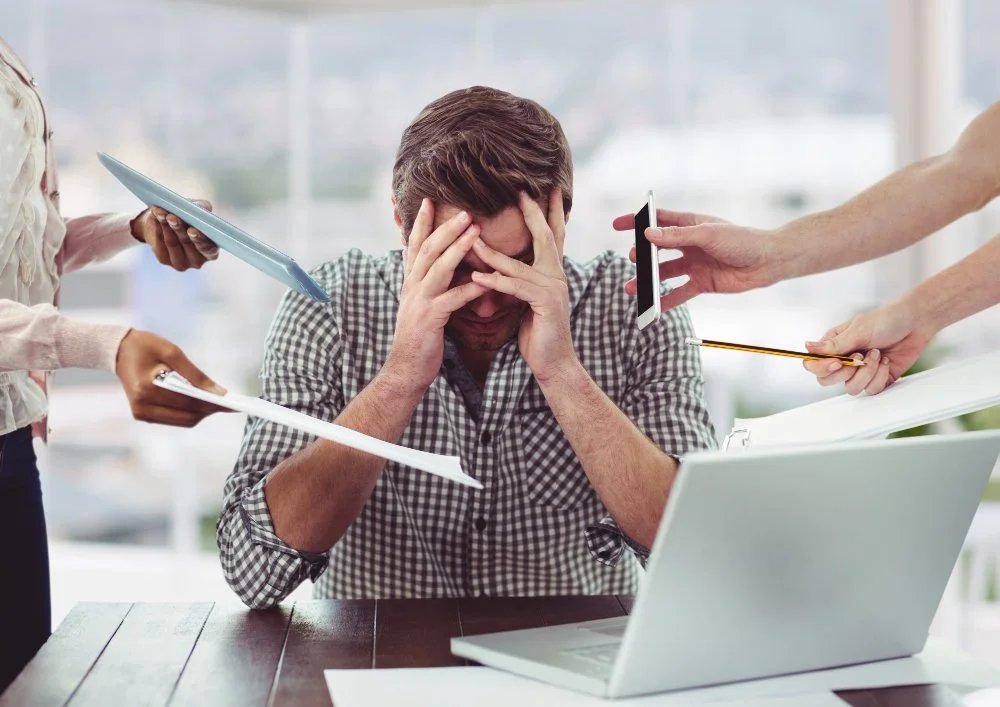Man sitting at a desk with a laptop, holding his head in frustration while multiple hands hold documents, a phone, a notebook, and a pencil towards him.