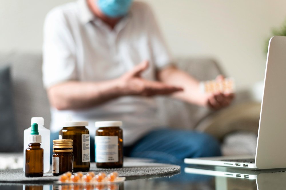 Person wearing a face mask sitting on a sofa with medication bottles and pills on a table in front of them, and a laptop nearby.