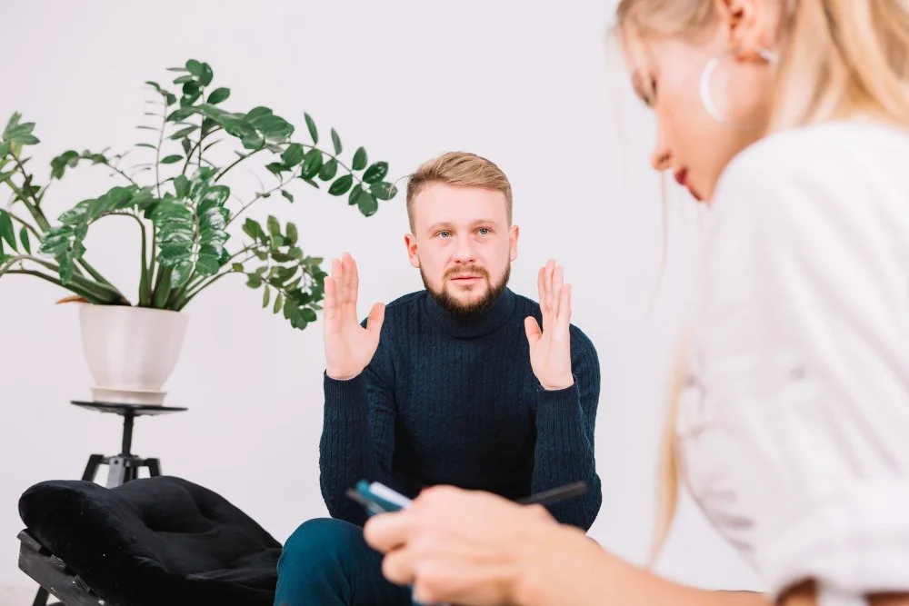 A man with a beard and wearing a dark sweater is gesturing with his hands during a therapy session while a woman in a white shirt is looking down at her phone.