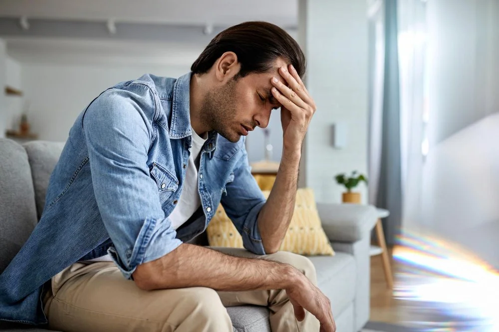 A man sitting on a couch with his head in his hand, appearing stressed or worried in a living room.
