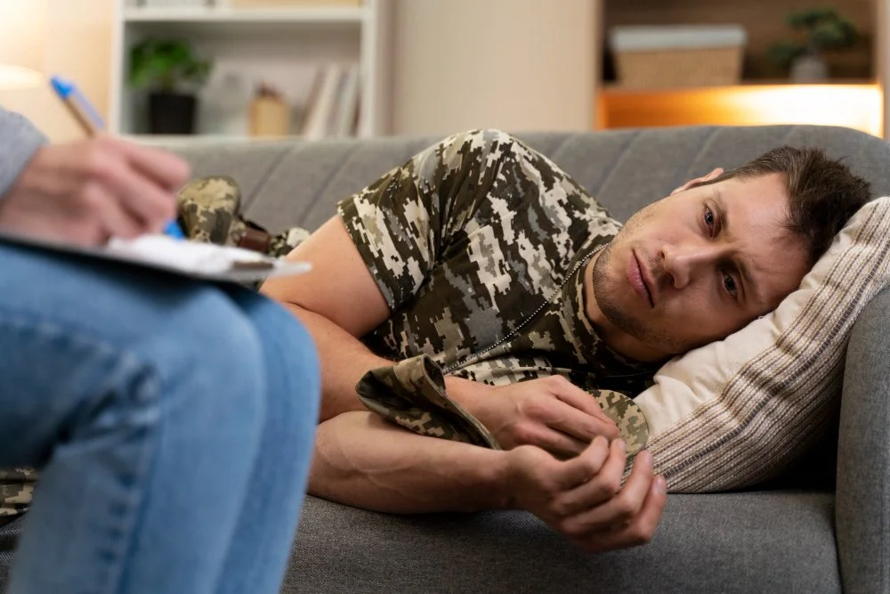 A man in military camouflage shirt lying on a gray couch, looking confused or concerned, while a person takes notes nearby.