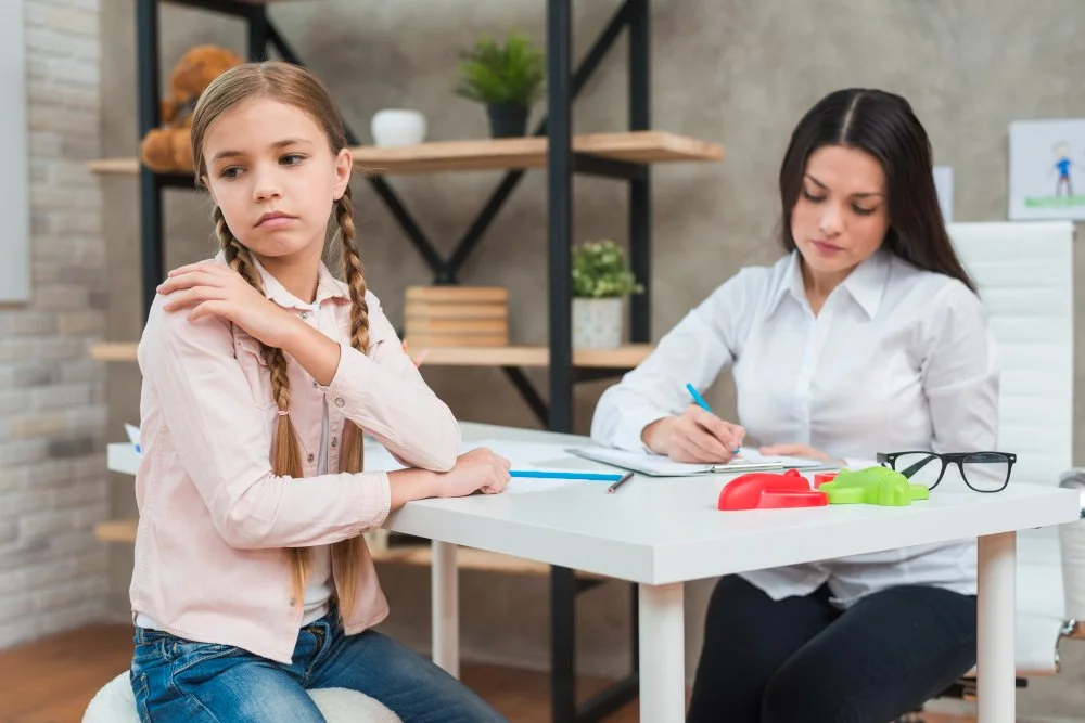 A young girl sitting at a therapy or counseling session with a woman, possibly a therapist, taking notes at a white table. The girl appears thoughtful, and the setting is a modern office with shelves, plants, and a brick wall in the background.