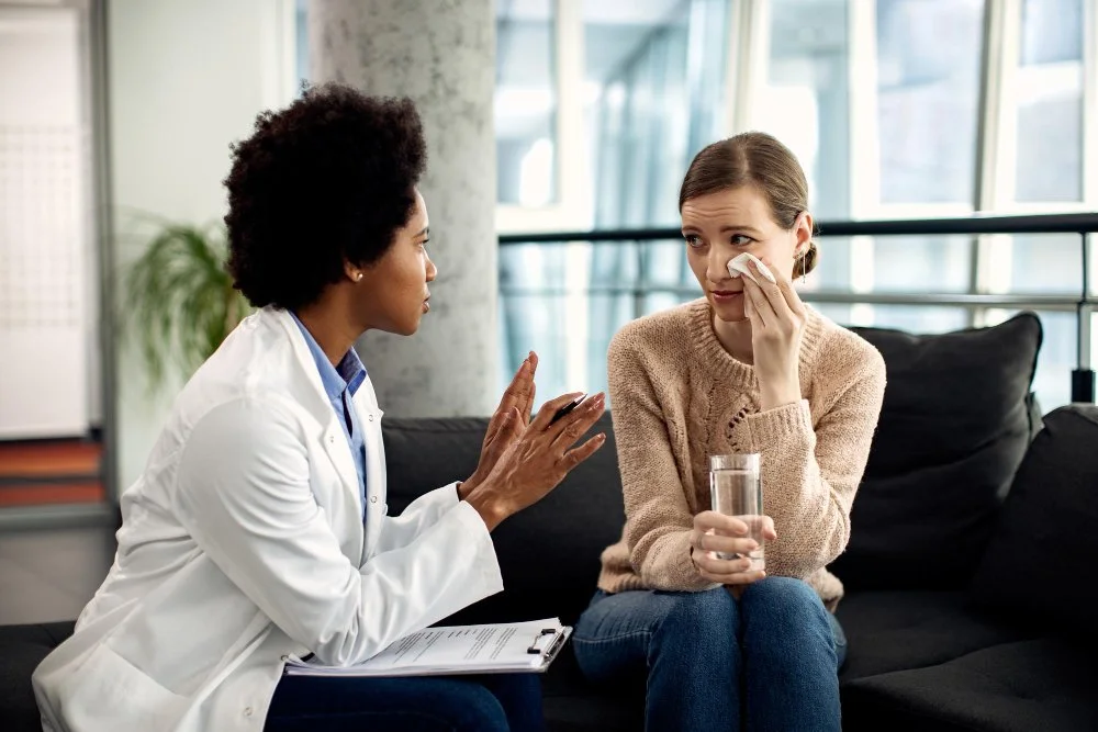 A female doctor talking to a woman who is holding a tissue to her nose, possibly indicating she is unwell. The woman is seated on a sofa with a glass of water in hand, in a modern indoor setting.