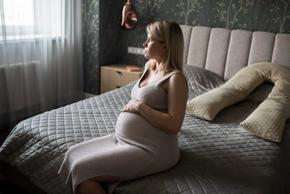 A pregnant woman sitting on a bed in a bedroom, looking out the window.