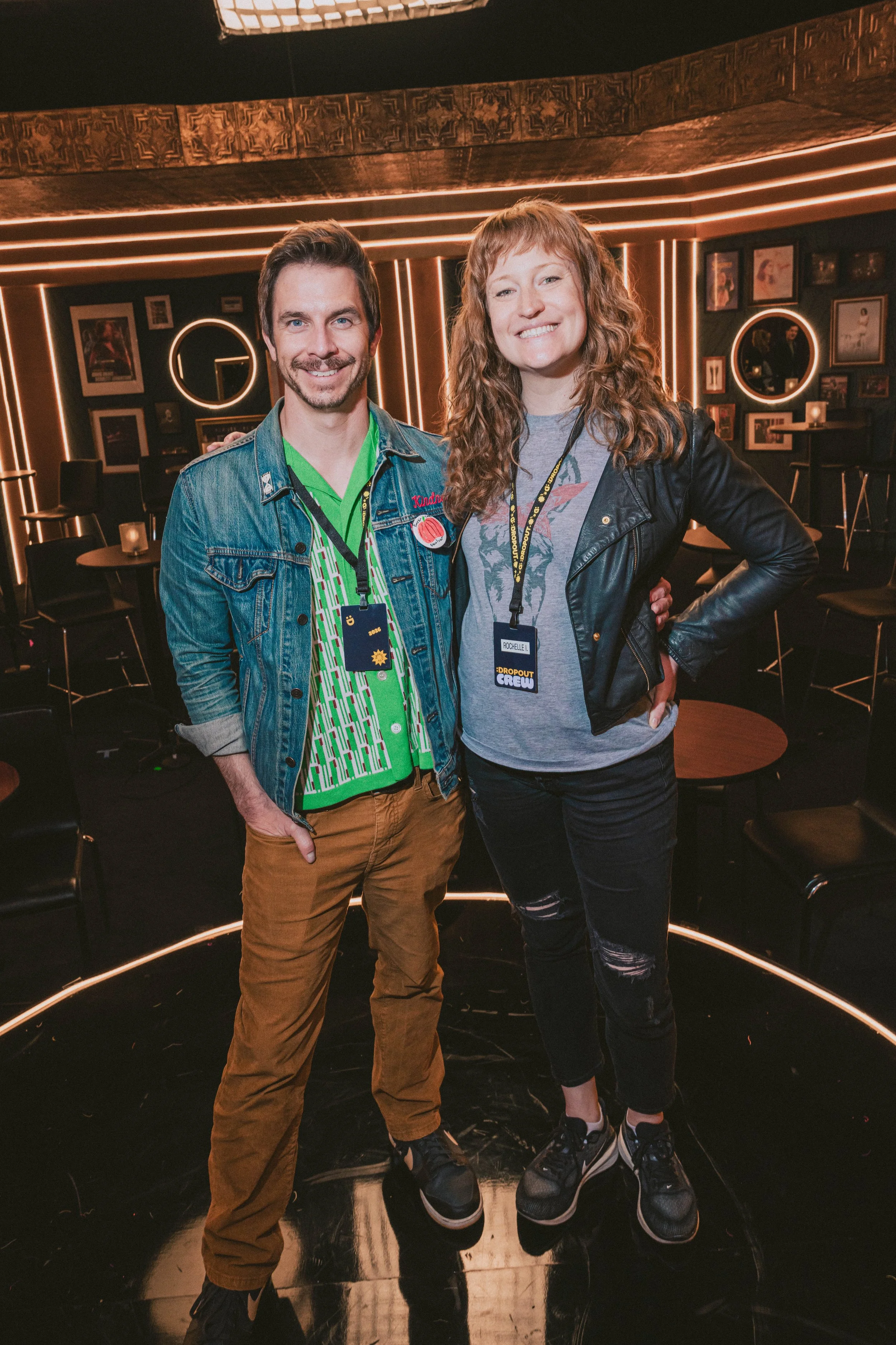 Two people standing together, smiling, in a dimly lit room with framed pictures on the walls and decorative lighting. The person on the left is a man with dark hair, wearing a denim jacket, green shirt, and brown pants. The person on the right is a woman with curly brown hair, wearing a black leather jacket, gray graphic T-shirt, ripped black jeans, and sneakers.