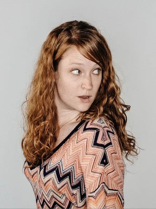 A woman with long, curly red hair looking to her right against a plain white background.