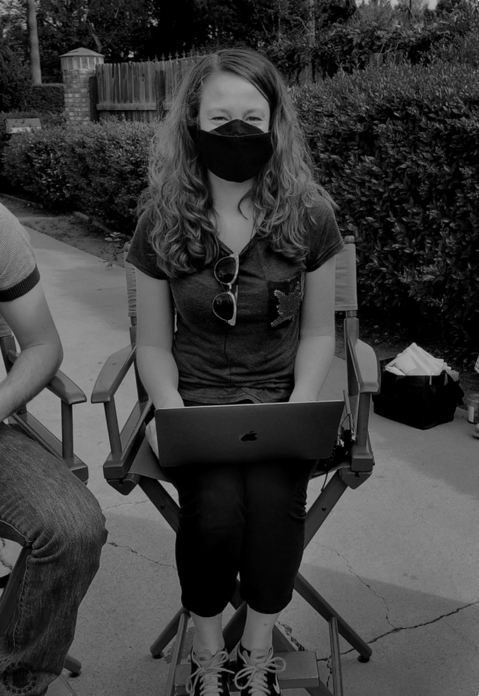 A young woman with curly hair sitting outside on a chair, wearing a face mask, sunglasses hanging from her shirt, and working on a laptop.