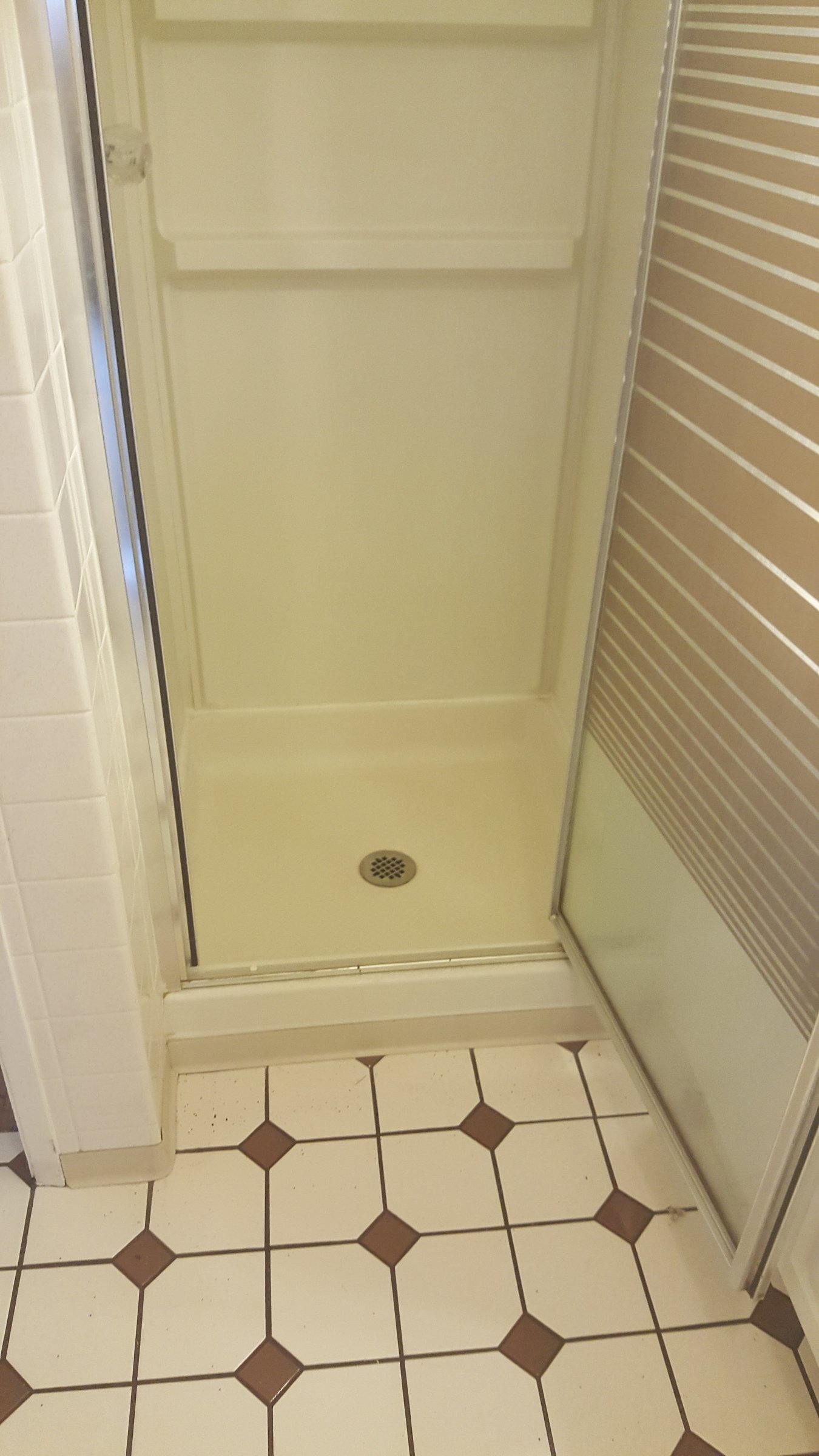 A shower stall with a brass drain cover, sliding glass door, beige tiles, and a brick-patterned wall.