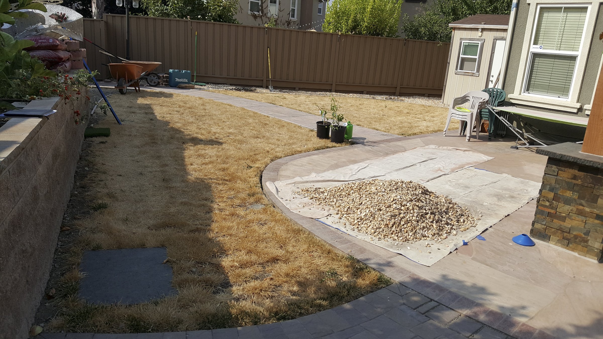 Backyard with a stone pathway, a small patch of dry grass, and gardening supplies. There are chairs stacked near a house, a fence, and some potted plants.