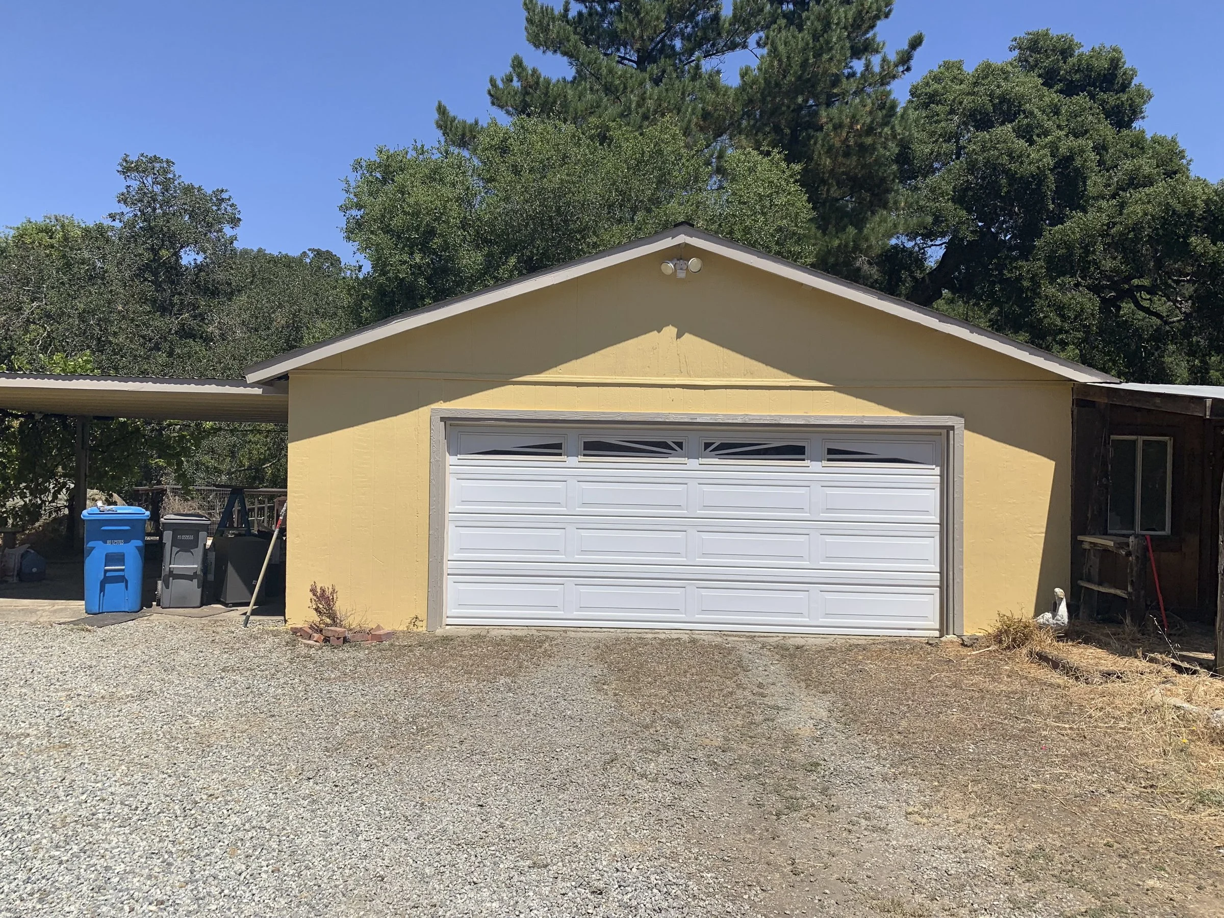 A yellow detached garage with a white roll-up door, a gravel driveway, trash bins to the left, and trees in the background under a clear blue sky.