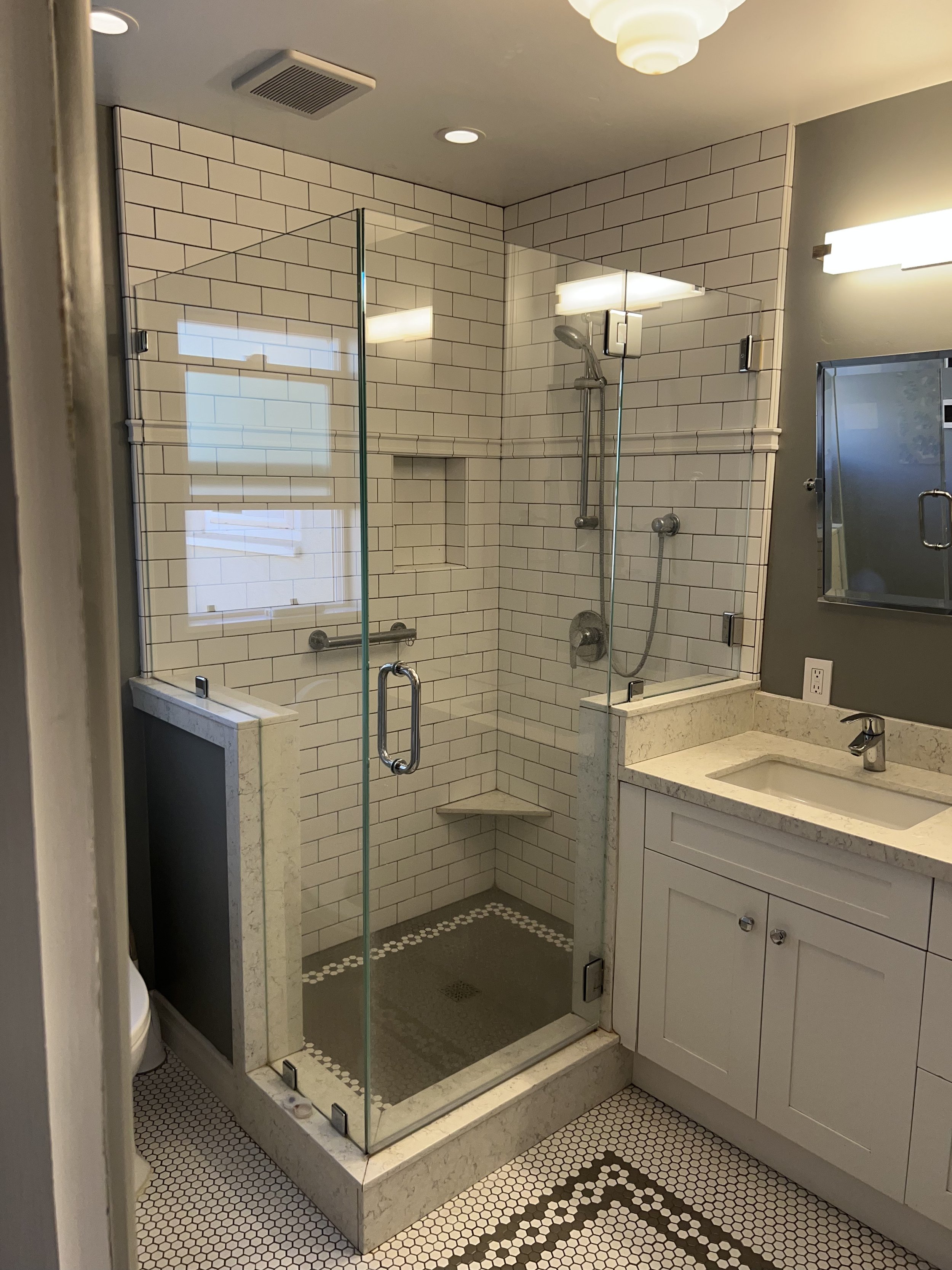 A bathroom with a walk-in shower enclosed by glass, white tiled walls, a gray vanity with a sink, and hexagonal tile flooring with a black and white pattern. There is a window, ceiling lights, and a mirror.