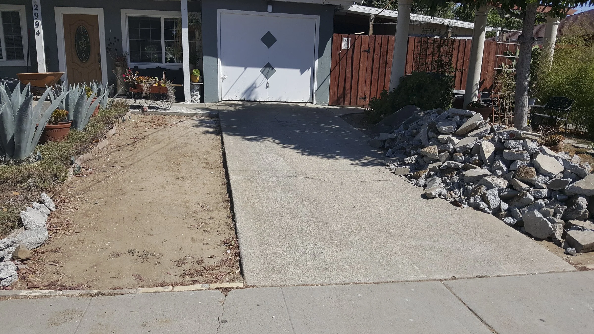 A residential driveway under construction with a pile of rocks on the right, an area of dirt on the left, and a finished concrete section in the middle. The house's front door and window are visible, with potted plants and a wooden fence.