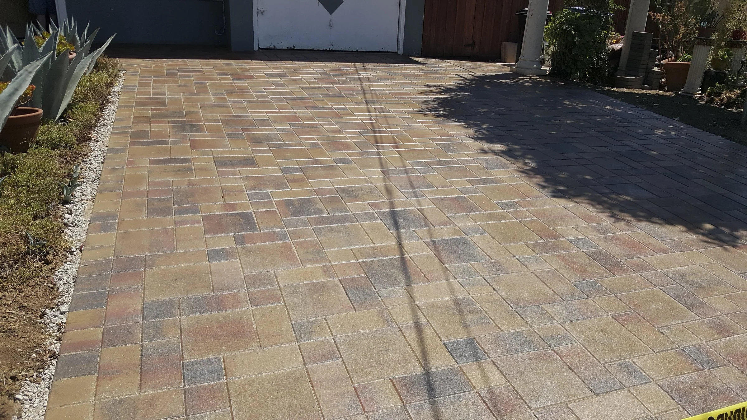 A newly installed brick patio with multicolored bricks in front of a garage with closed white doors, side garden beds with plants, and some shadows on the ground.