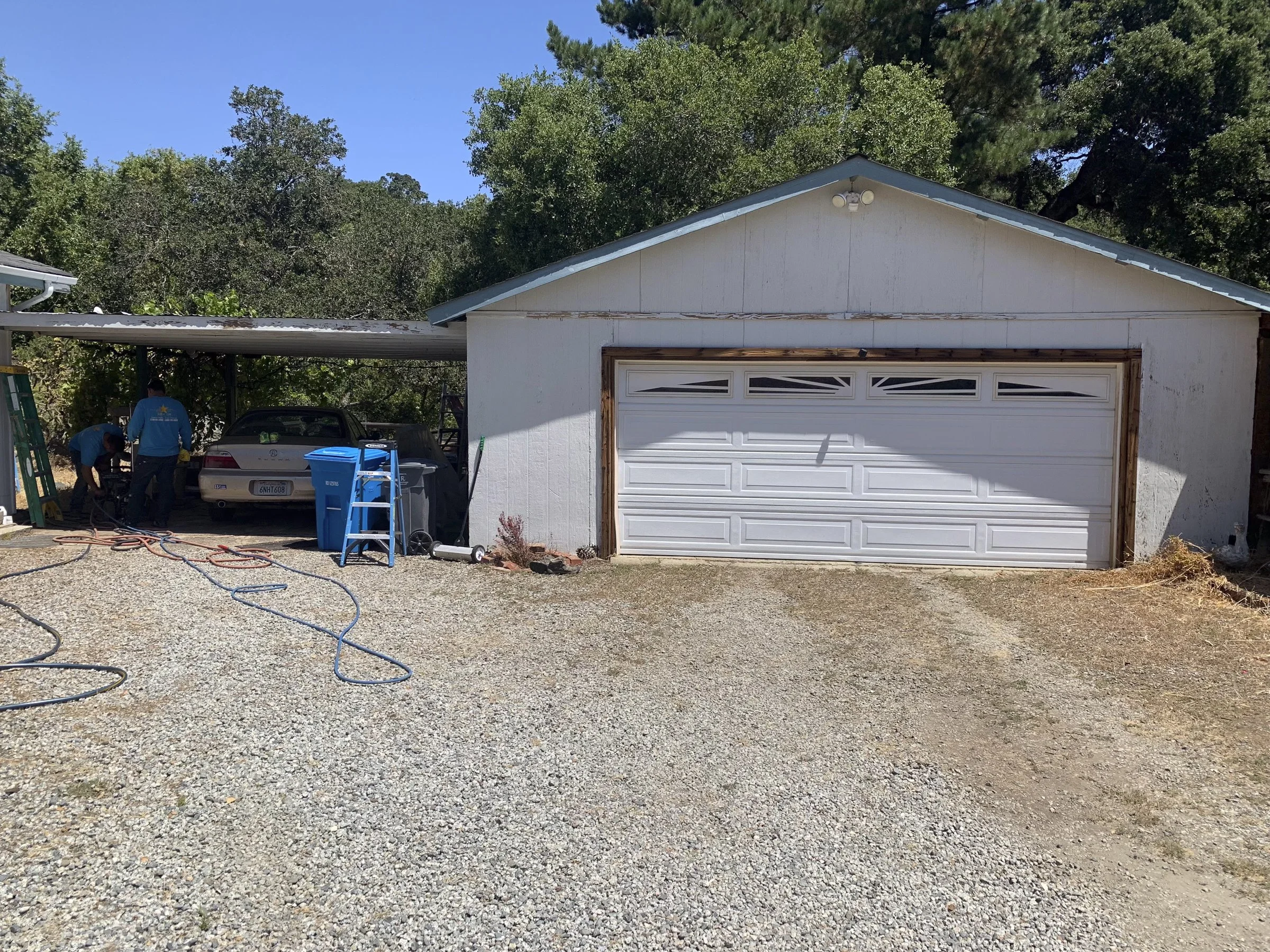 A garage with a partially installed garage door, two workers working nearby, and various tools and equipment around, with trees and clear blue sky in background.