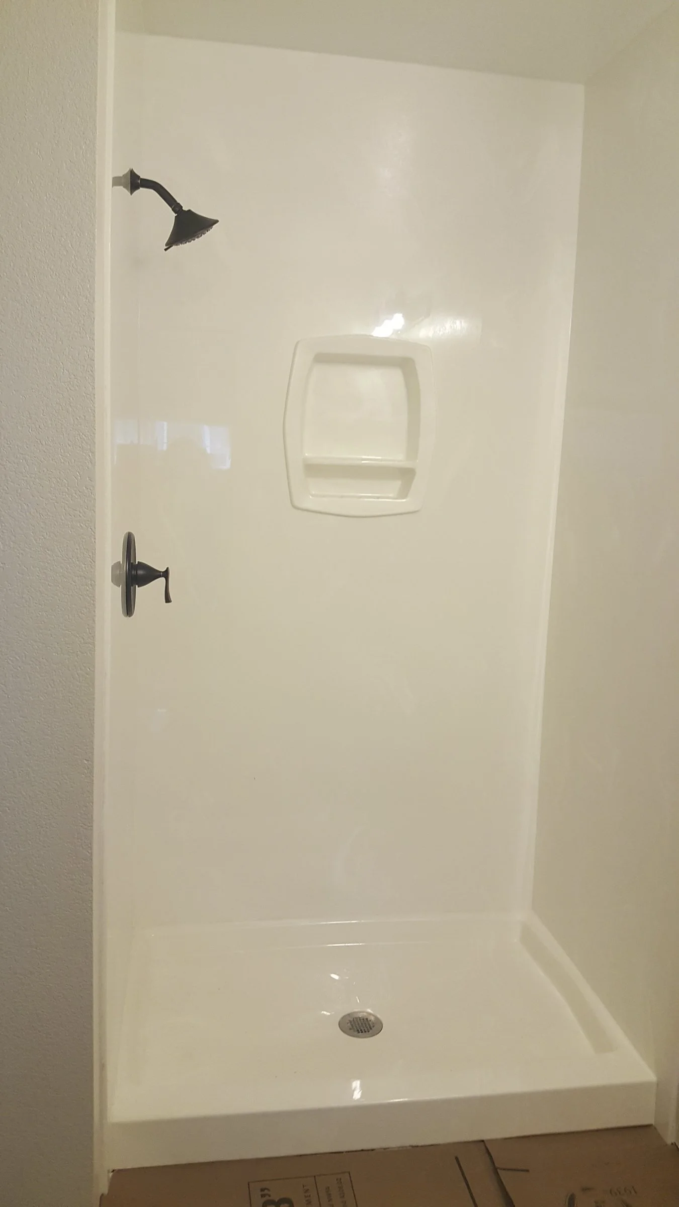 A plain white shower stall with a black showerhead, a black handle, and a built-in shelf. The shower has a drain at the bottom and is surrounded by a beige wall.