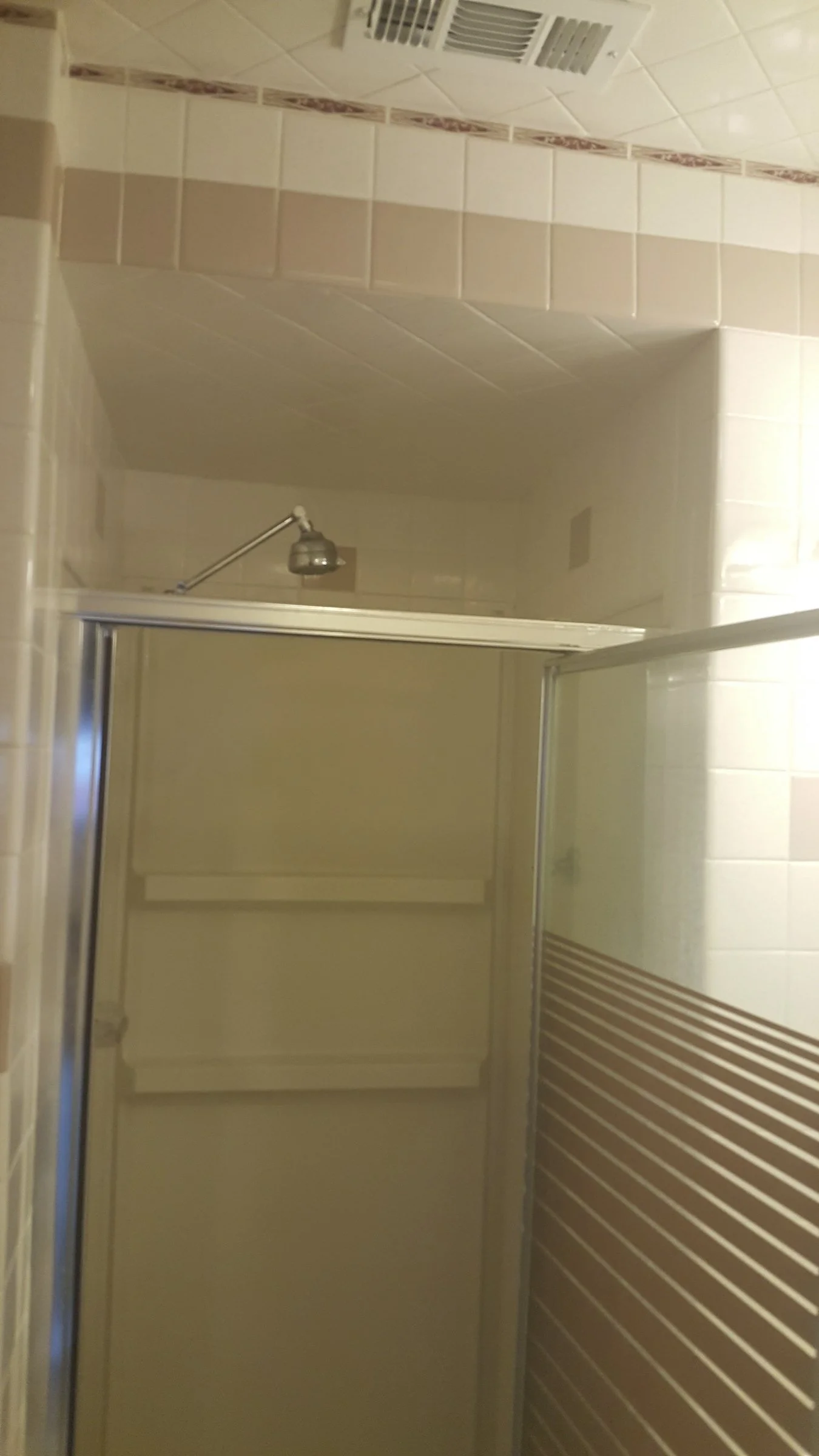 Bathroom with beige and tan tiled walls, shower area with chrome door frame, shower head, and wall vent.