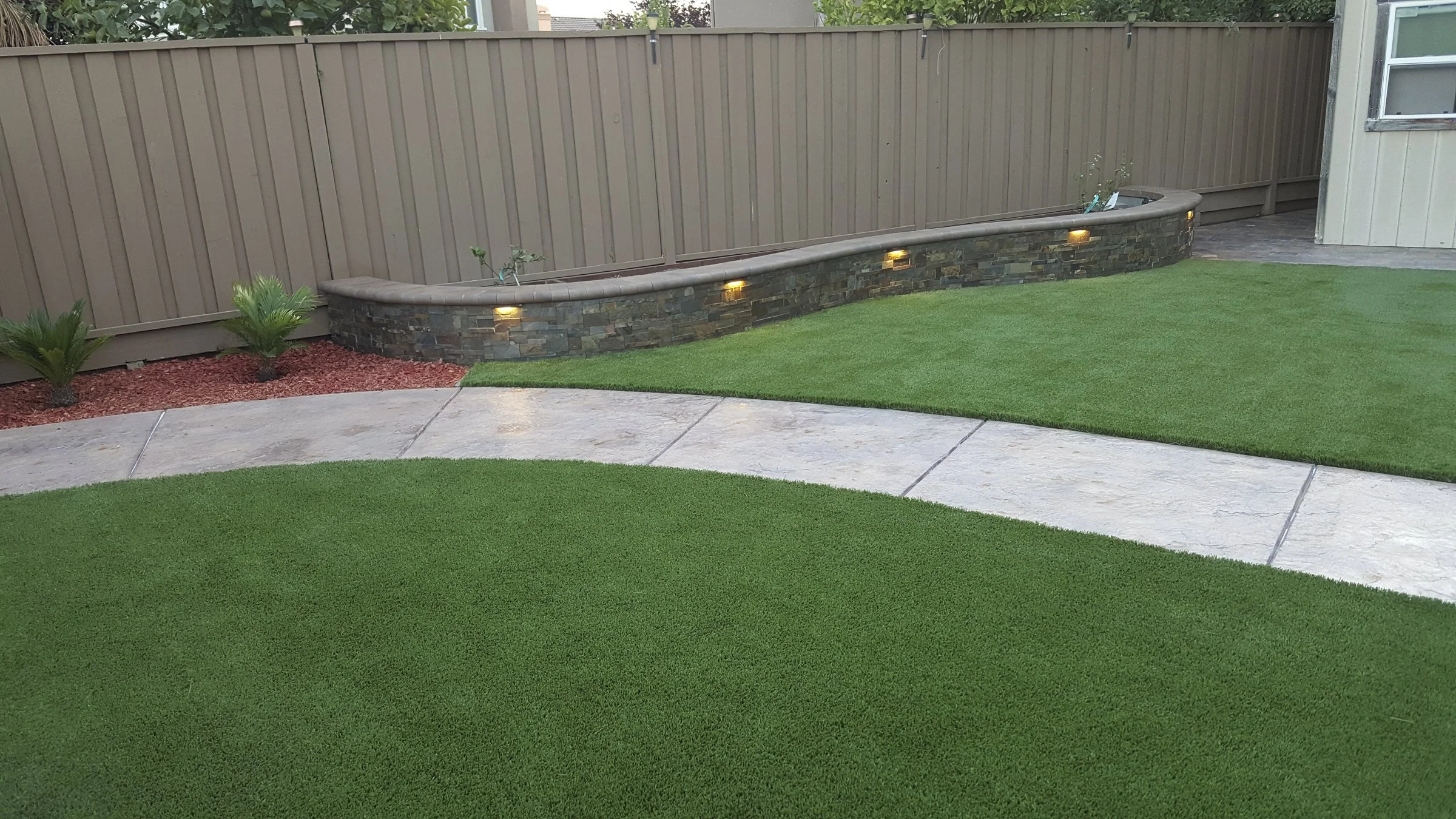 A backyard with a curved stone retaining wall lit with small lights, a wooden privacy fence, green grass, small palms, and a concrete pathway.