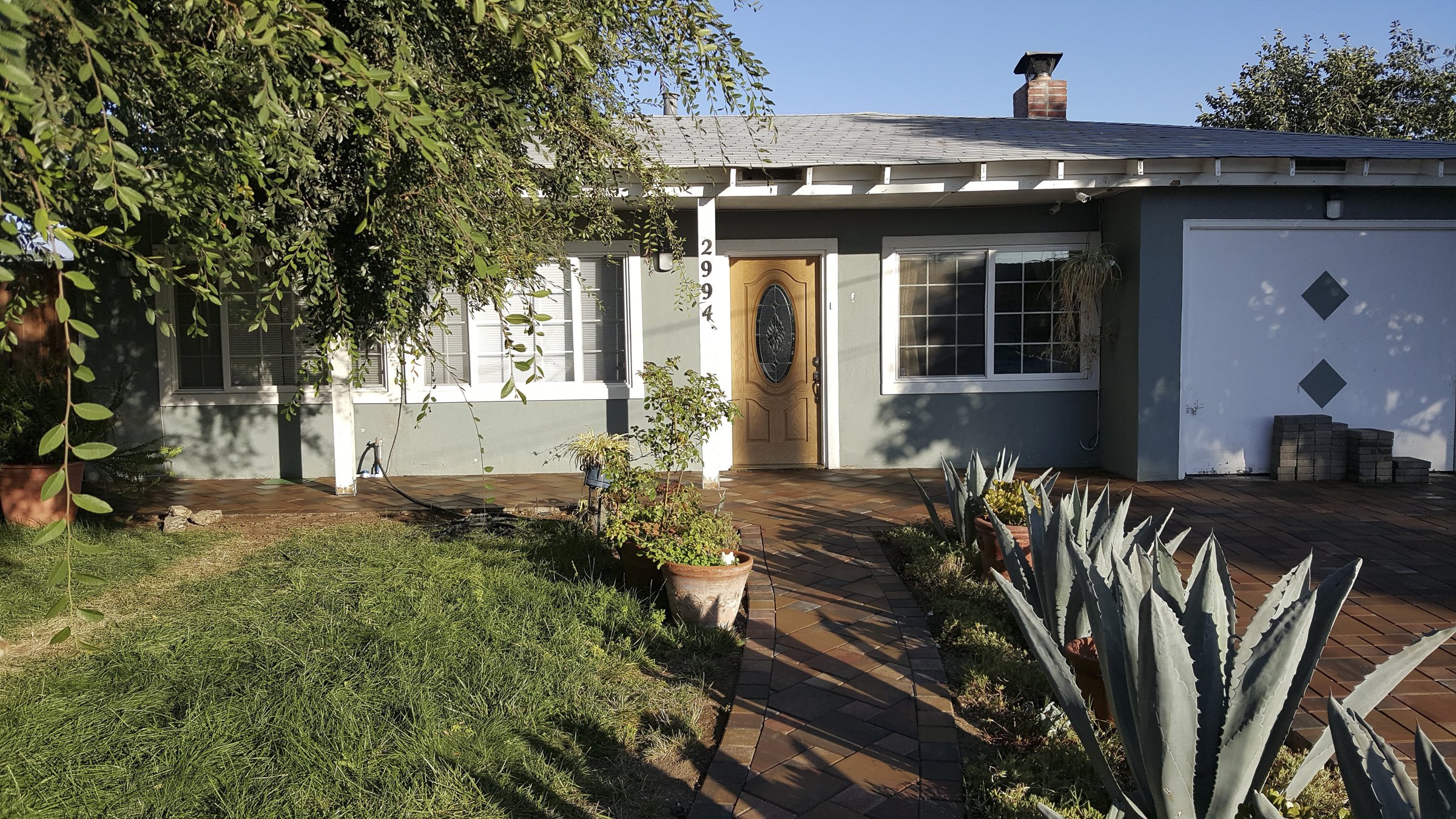 A house with a front yard featuring a brick walkway, potted plants, and a desert plant in the garden. The house has a gray exterior with a wooden front door and white window frames. A large tree partially shades the yard, and a garage with two diamond-shaped decorations is visible on the right.