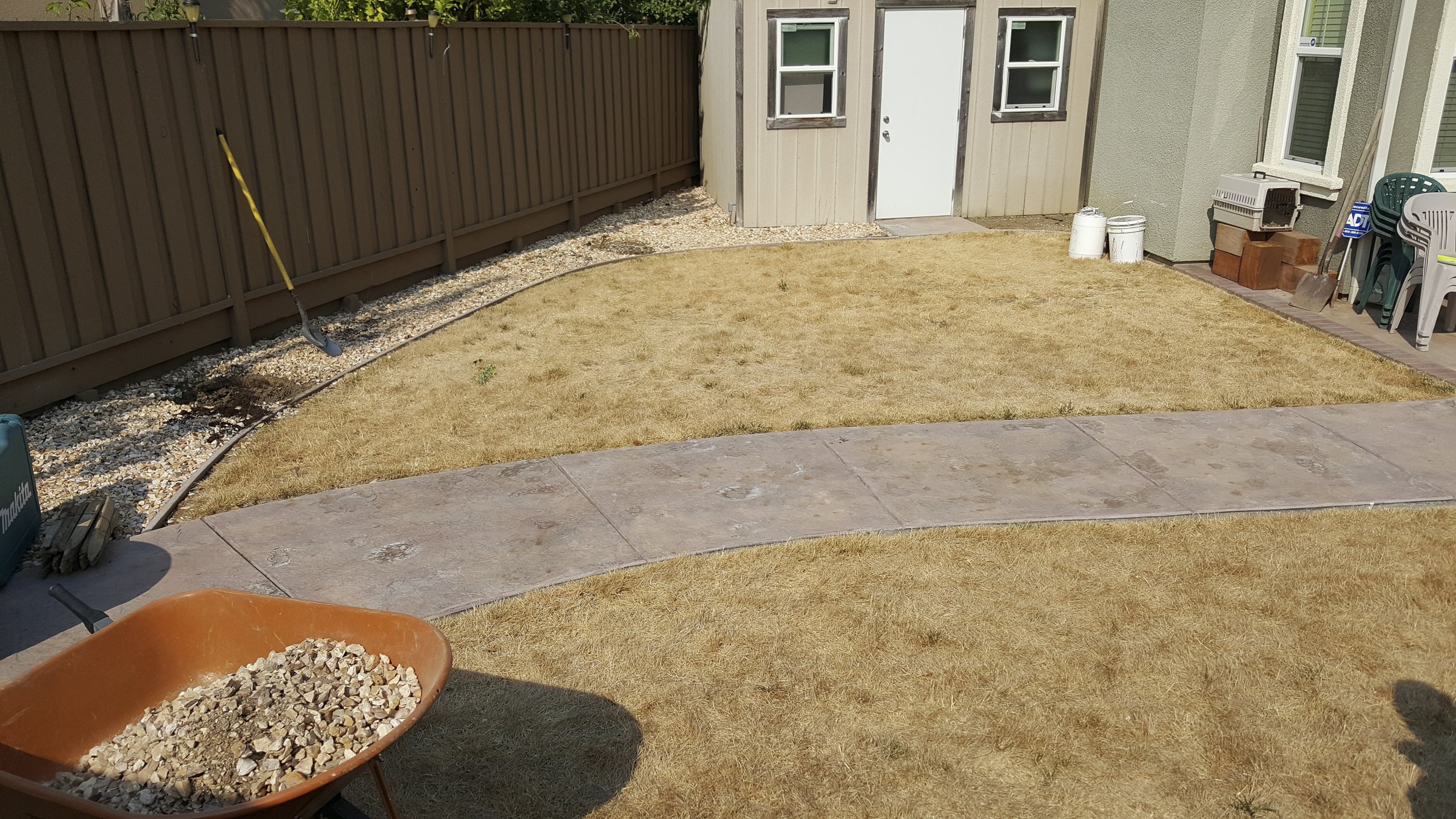 Backyard with a brown wooden fence, a small shed with a white door, and a patio area with stacked chairs, buckets, and a wheelbarrow filled with gravel.