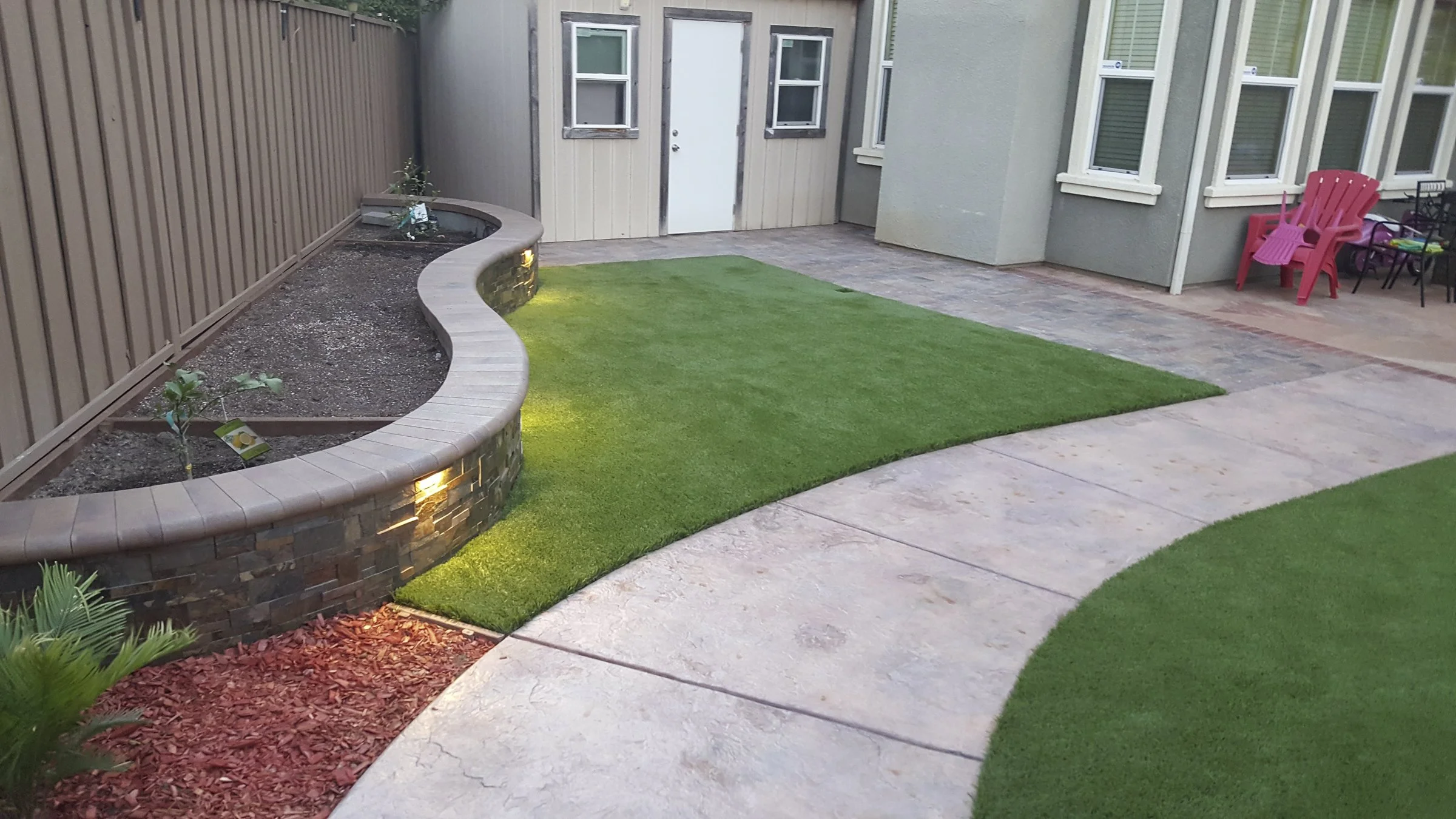 Backyard with a curved brick-edged flower bed, artificial grass, and a concrete patio. There are chairs and children's toys near the house.