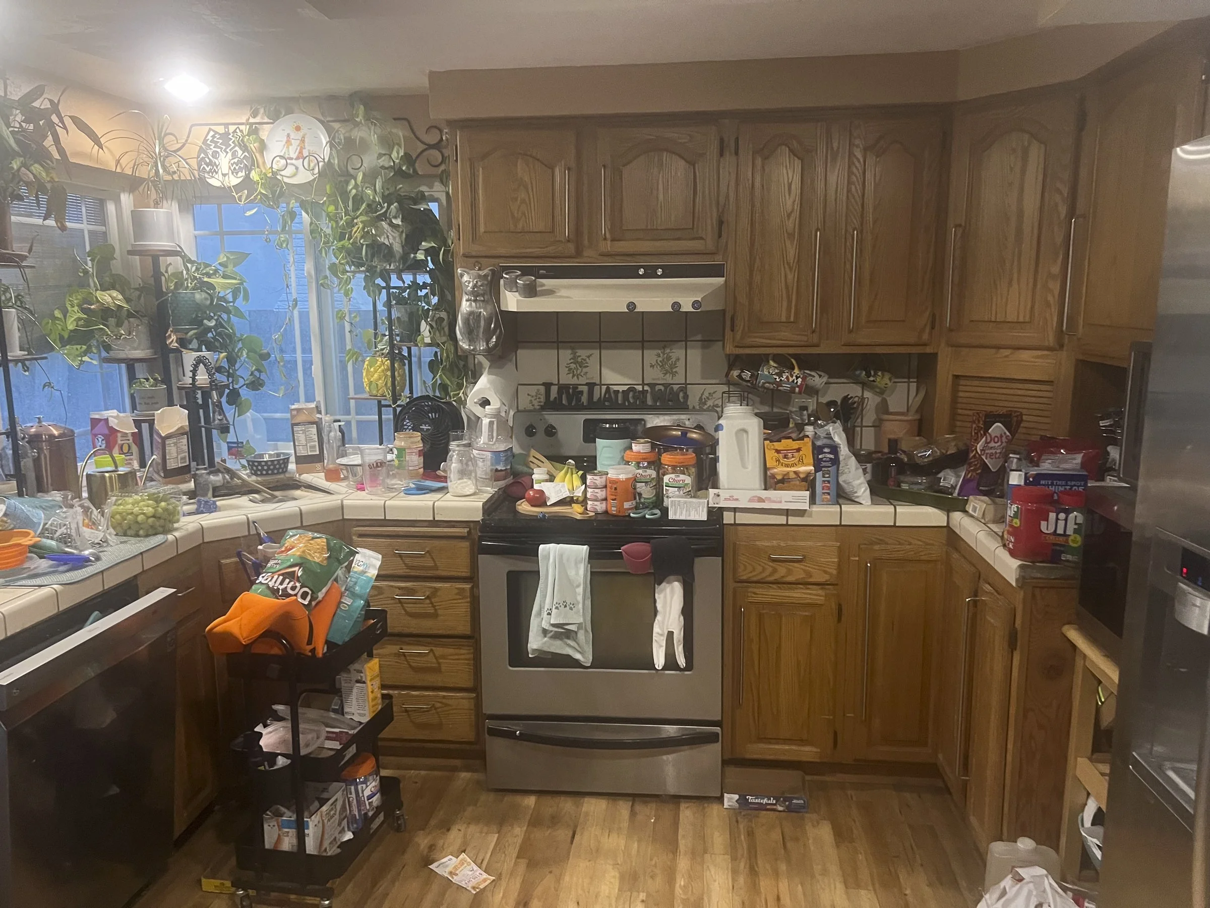 Cluttered kitchen with wooden cabinets, a stove, and various food items and dishes on the counter.