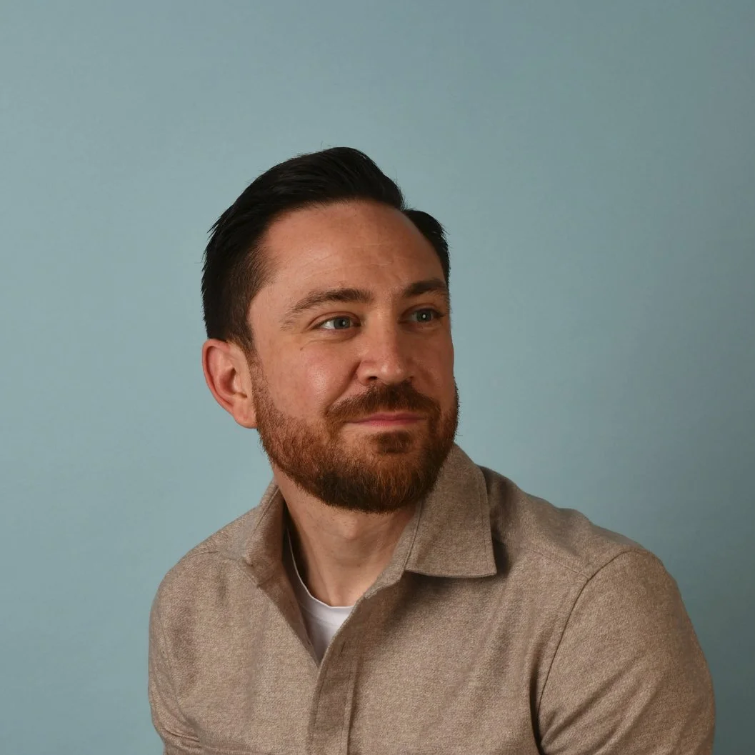 A man with dark hair, a beard, and light skin, wearing a light brown shirt, looking to the side against a plain, light blue background.