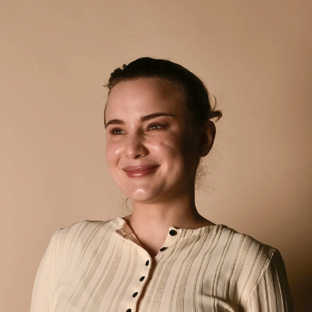 A woman smiling with her head slightly tilted to the left, wearing a beige shirt with black buttons on a plain beige background.