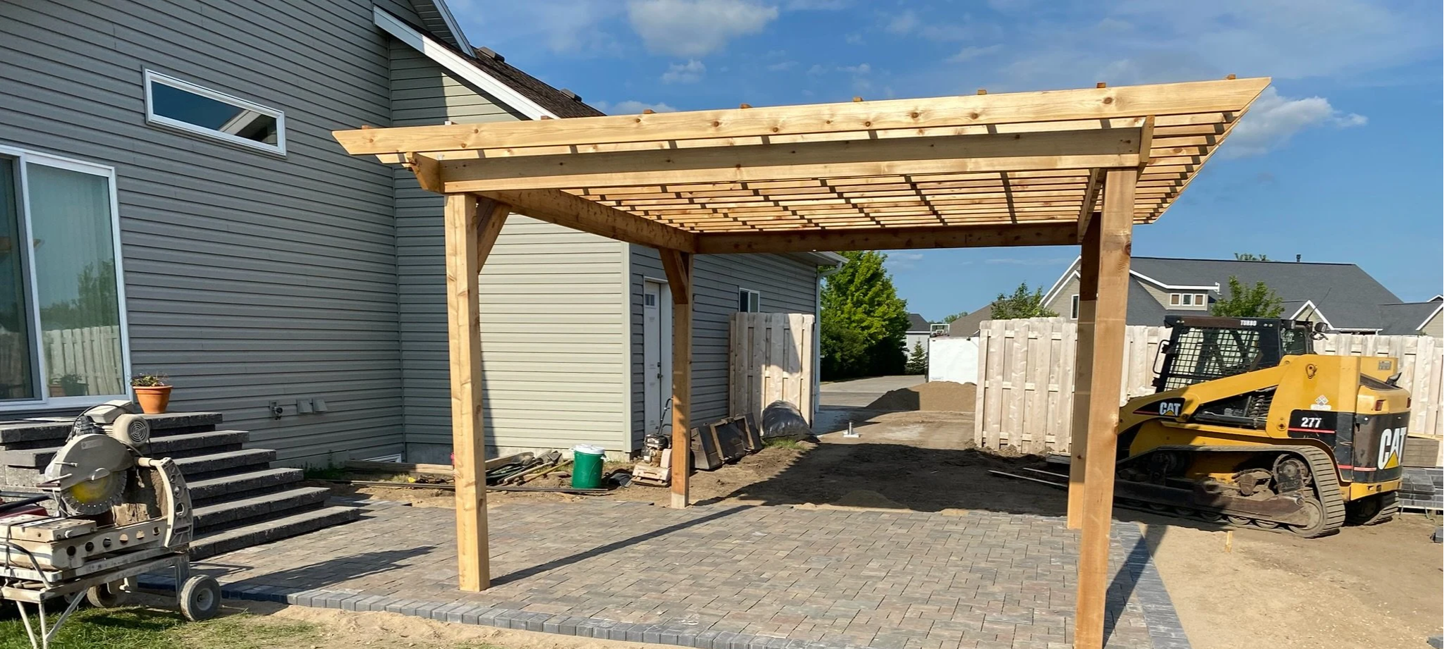 Construction site with a wooden carport structure, brick patio, and construction equipment, with neighboring houses and a clear blue sky.