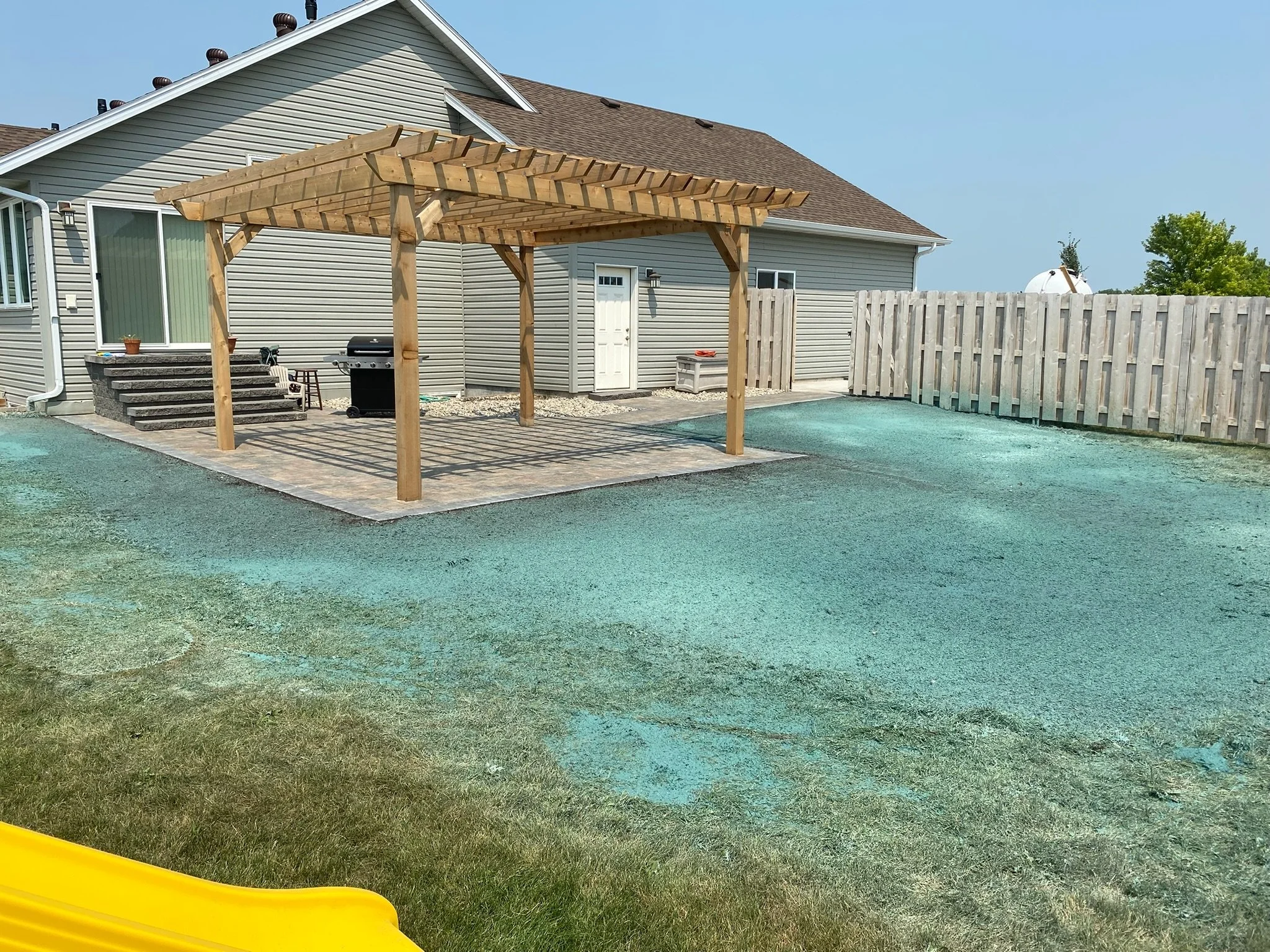 Backyard patio with a wooden pergola, a barbecue grill, and a small staircase leading up to the house. There is a porch with potted plants and a sliding glass door. The yard has a mix of gravel and grass, fenced with wooden fencing, and a cloudy blue sky.