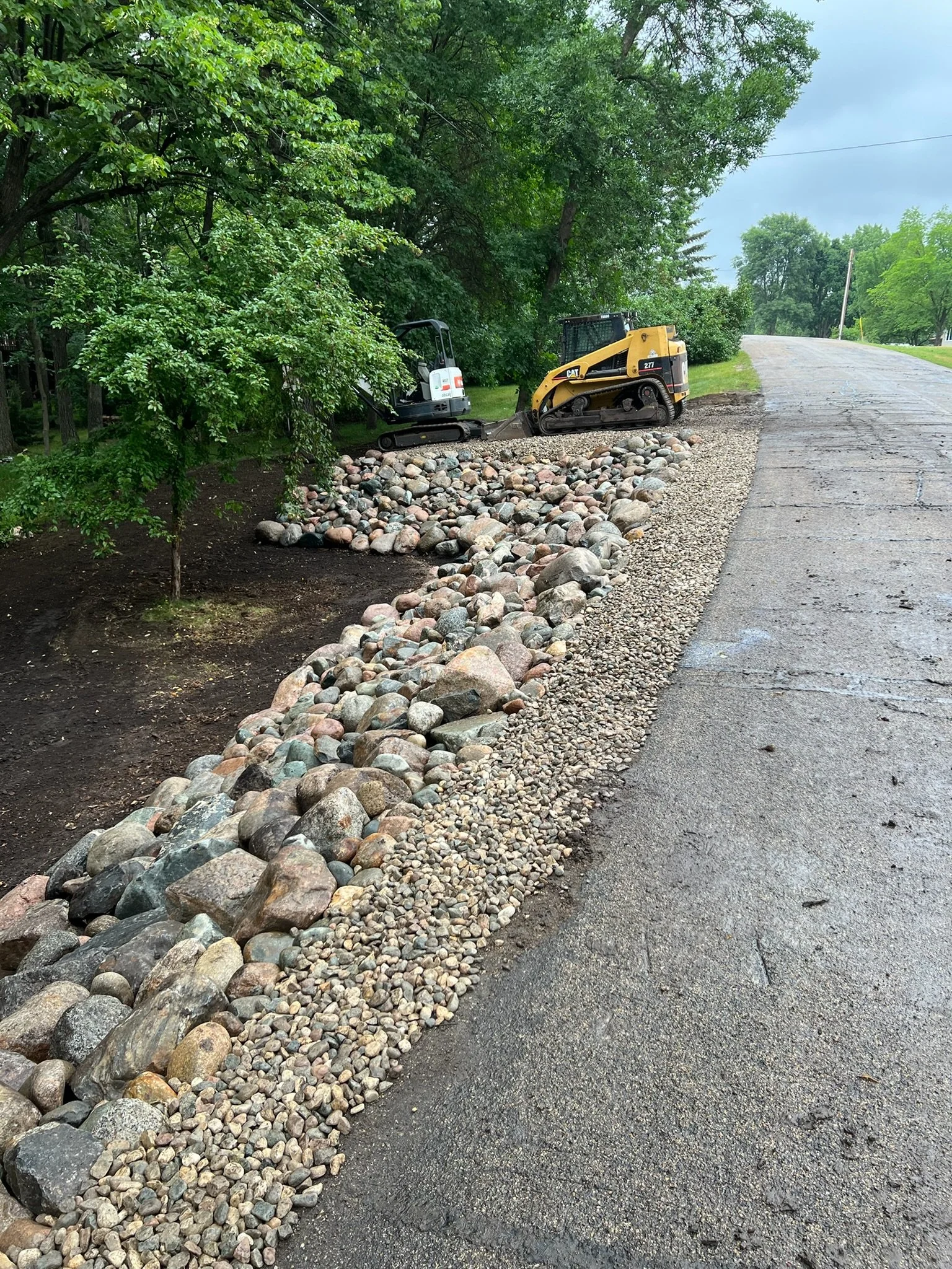 Construction equipment, including a mini excavator and a skid-steer loader, working on a roadside with gravel and rocks, and trees in the background.