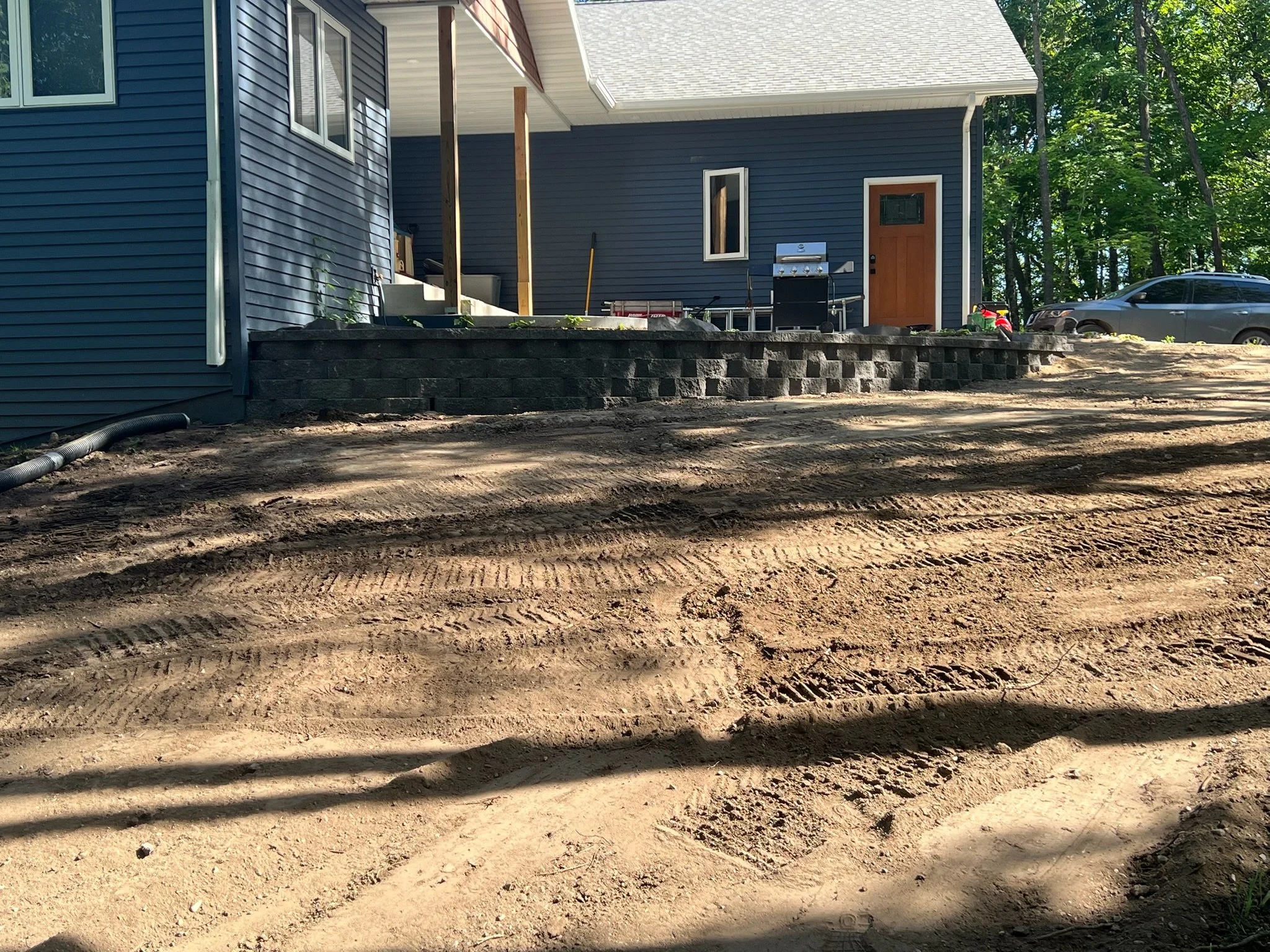 A house with dark blue siding and white trim, a wooden front door, and an outdoor patio with a grill, toys, and a ladder. A car is parked on the gravel driveway, and trees are in the background. The ground in the foreground is uneven dirt.