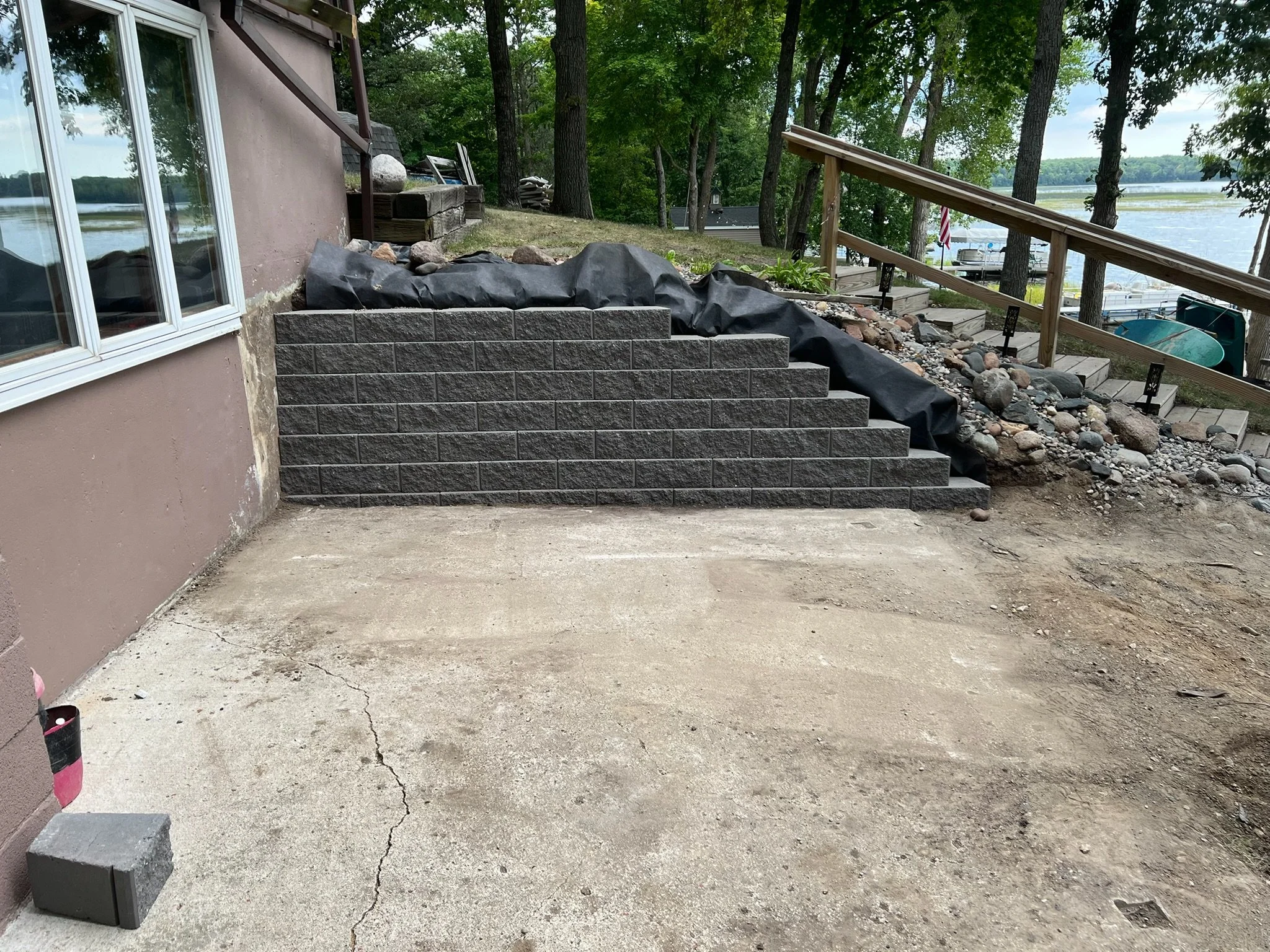A backyard with a concrete patio in the foreground, and a set of dark gray brick stairs leading up to the yard, which has rocks, dirt, and plants. A wooden railing runs along the yard's edge, overlooking a river with boats and trees on the other side.