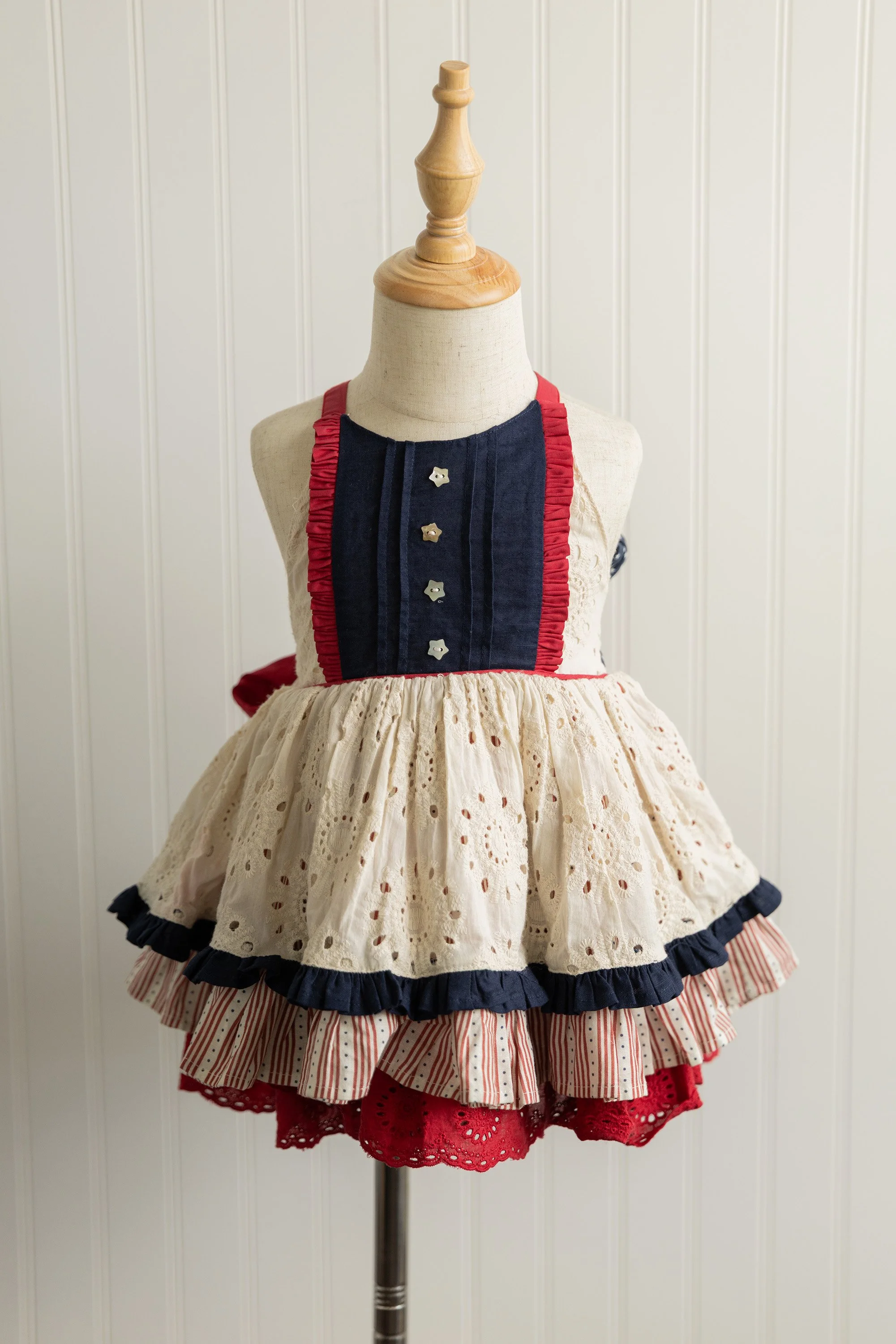 A dress displayed on a mannequin with a beaded bodice, ruffled collar, and a layered skirt featuring eyelet and striped patterns, set against a white wood-paneled background.
