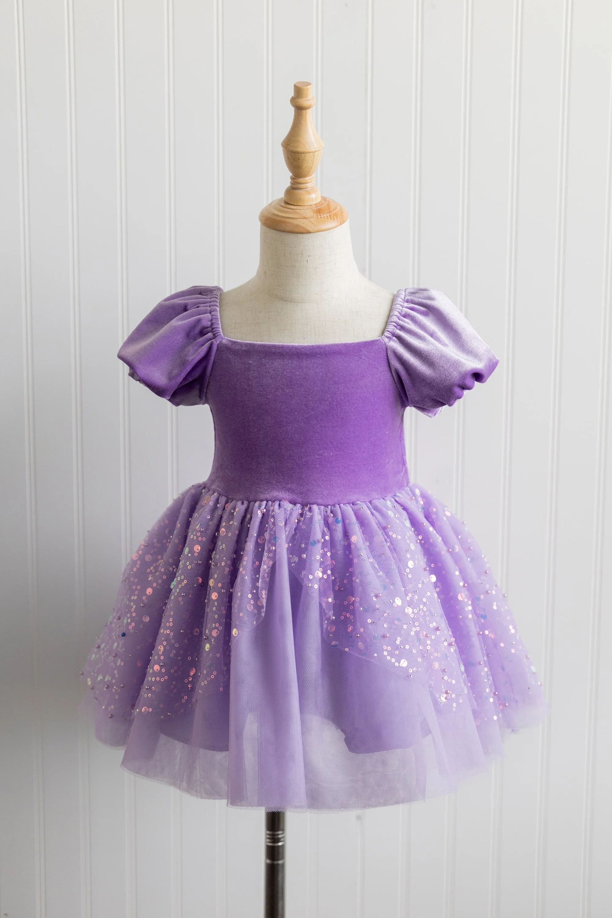 A children's dress on a mannequin, purple with puffed sleeves, a fitted bodice, and a glittery, tulle skirt, displayed against a white paneled wall.