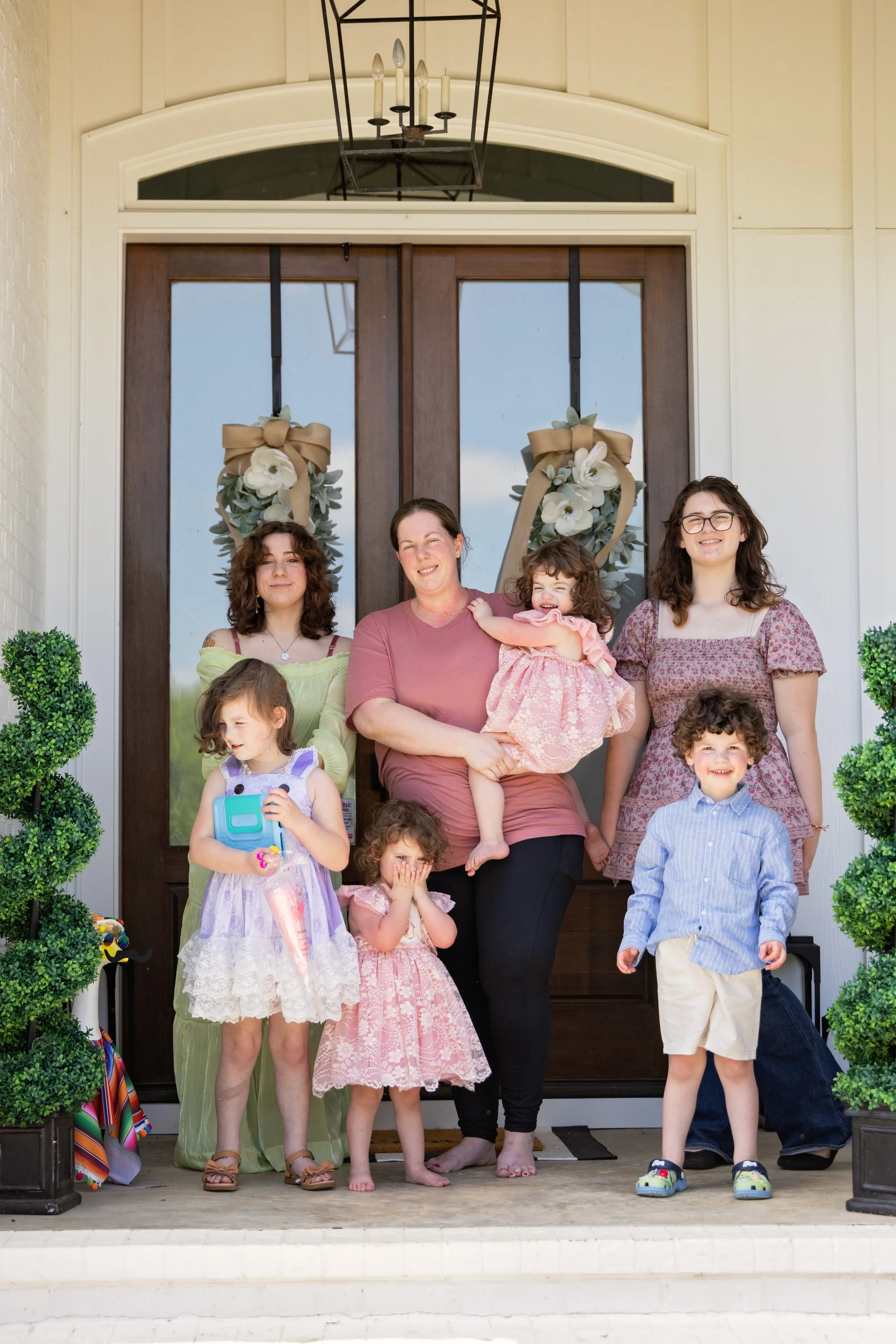 Family group standing on the porch of a house in front of a decorative front door with wreaths, flanked by two potted bushes, during daytime.