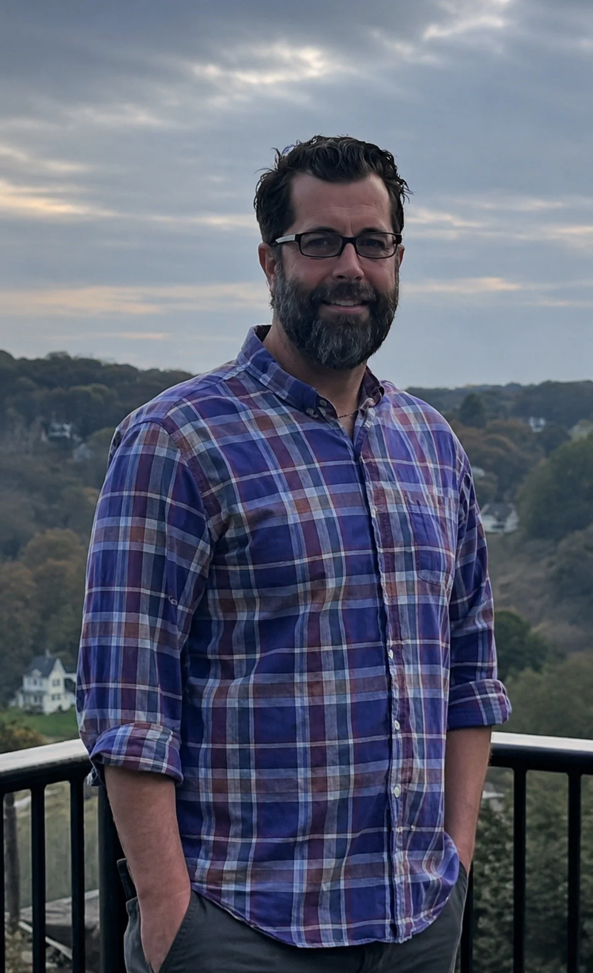 A man with dark hair, glasses, and a beard, wearing a plaid shirt, standing outdoors on a balcony with a scenic background of rolling hills, trees, houses, and a cloudy sky.