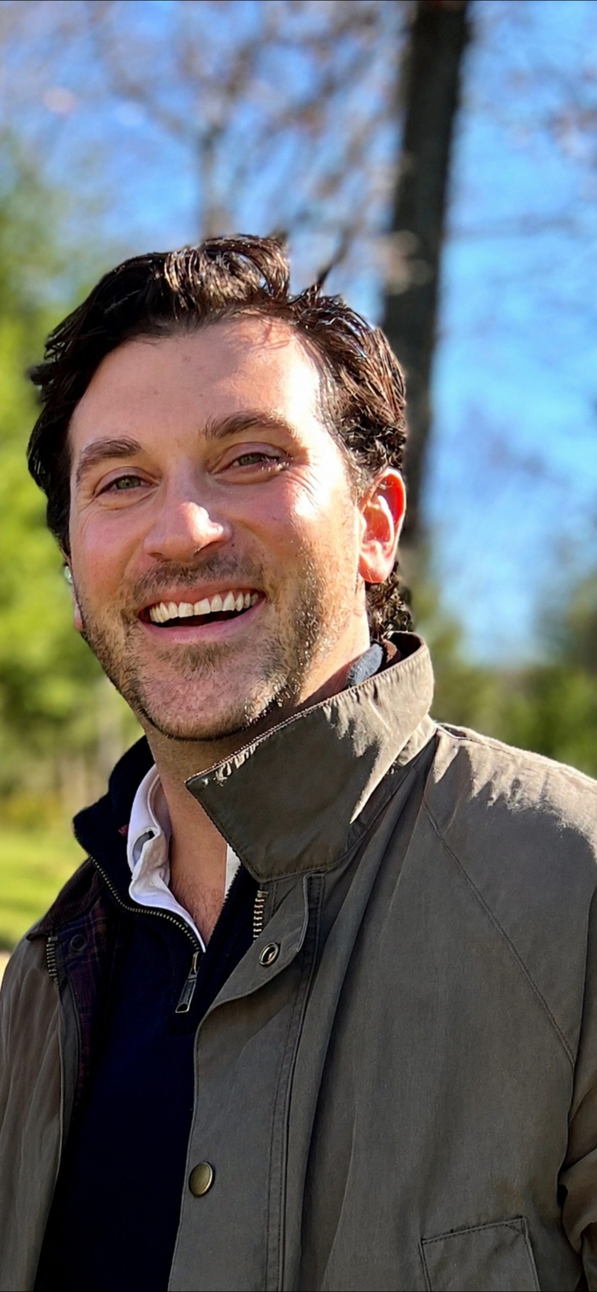 A smiling man outdoors with a tree and blue sky in the background, wearing a gray jacket and a white shirt.