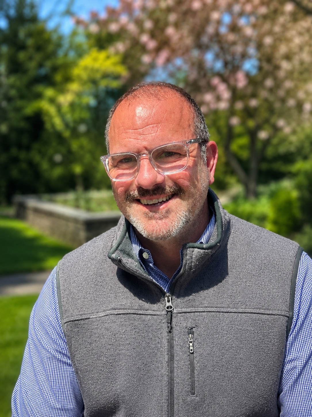 Smiling man with glasses outdoors with blooming pink flowers and greenery in the background.