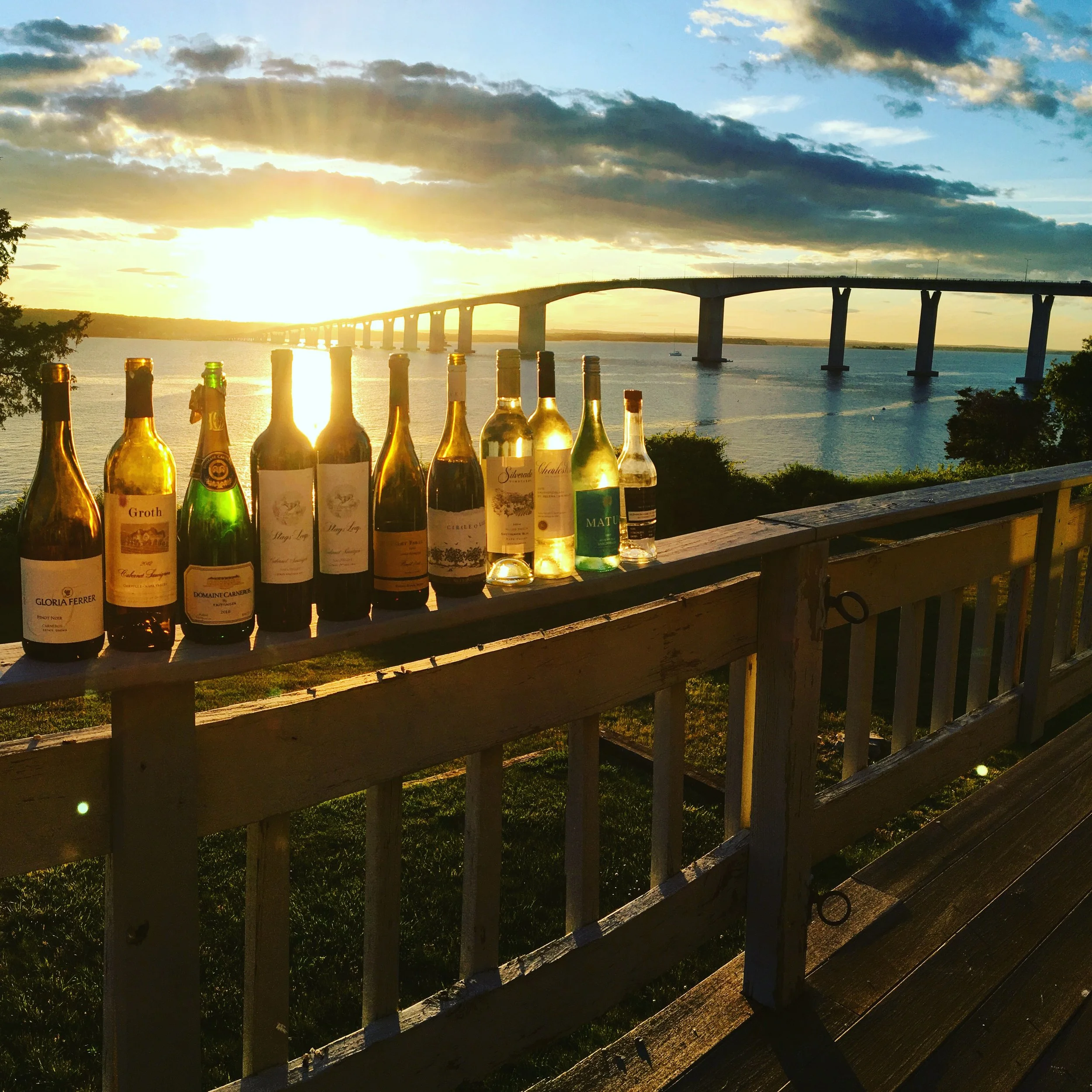 Sunset over a large body of water with a bridge stretching across it, viewed from a wooden deck with bottles lined up on the railing.