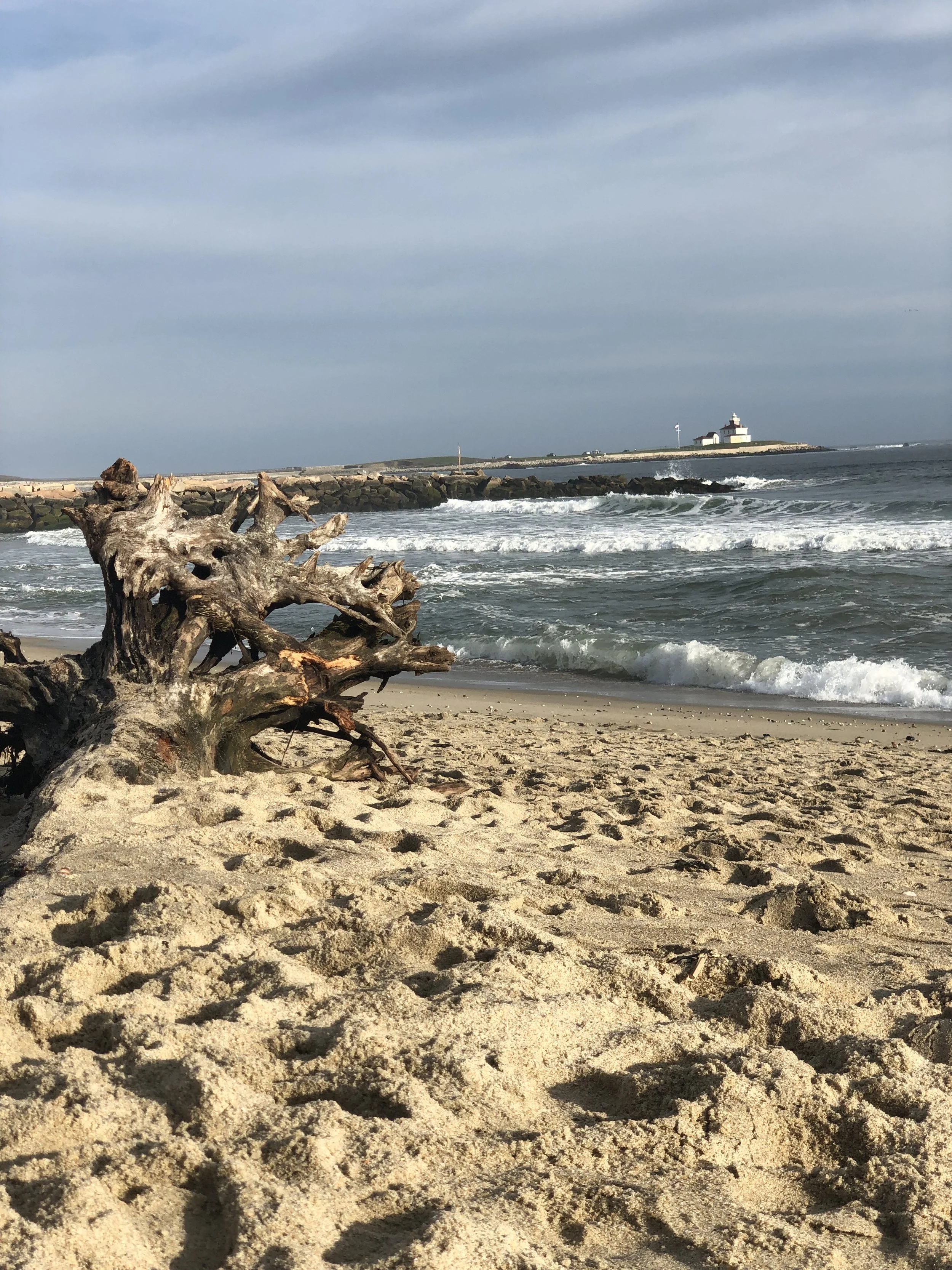 A sandy beach with a driftwood log in the foreground, ocean waves crashing onto the shore, a rocky jetty extending into the water, and a lighthouse on the distant point under a cloudy sky.